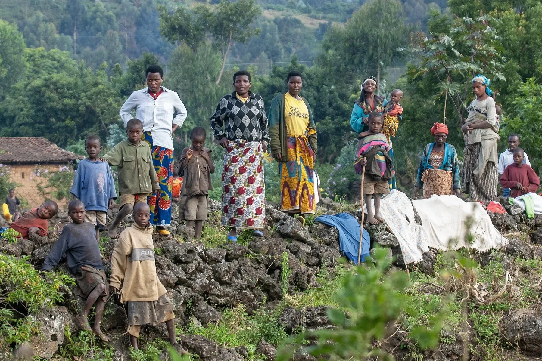 Group of women and children standing on a rocky hillside in a rural area with trees and a house in the background.