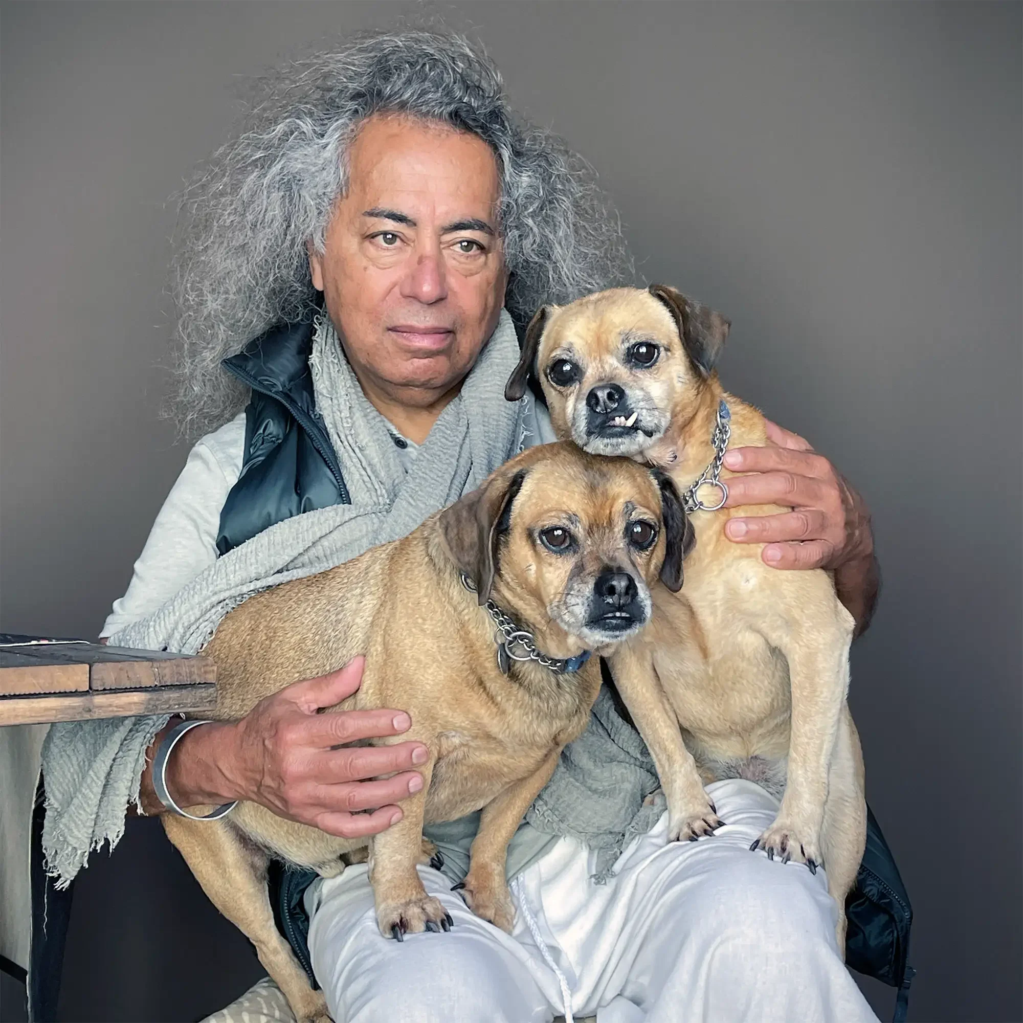 A man with curly gray hair wearing a gray shirt, a beige scarf, and a blue vest, sitting with two tan-colored dogs on his lap. The man looks directly at the camera with a neutral expression. The dogs, one with a short snout and a toothy smile, and th