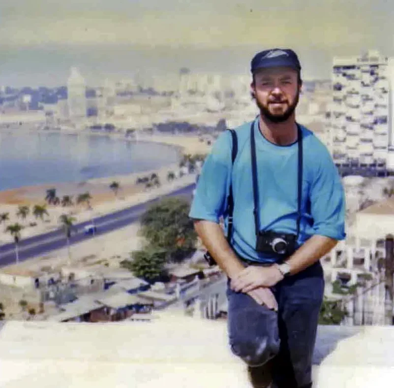 A man with a beard wearing a blue cap and t-shirt, carrying a camera around his neck, standing on a rooftop with a cityscape, a body of water, and palm trees in the background.