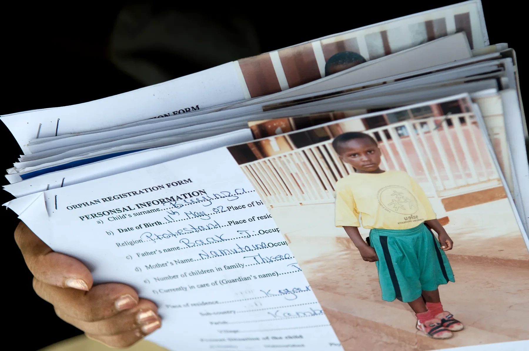 A hand holding a stack of orphan registration forms and photographs of children, including a young boy in a yellow shirt and green shorts standing outdoors near a fence.