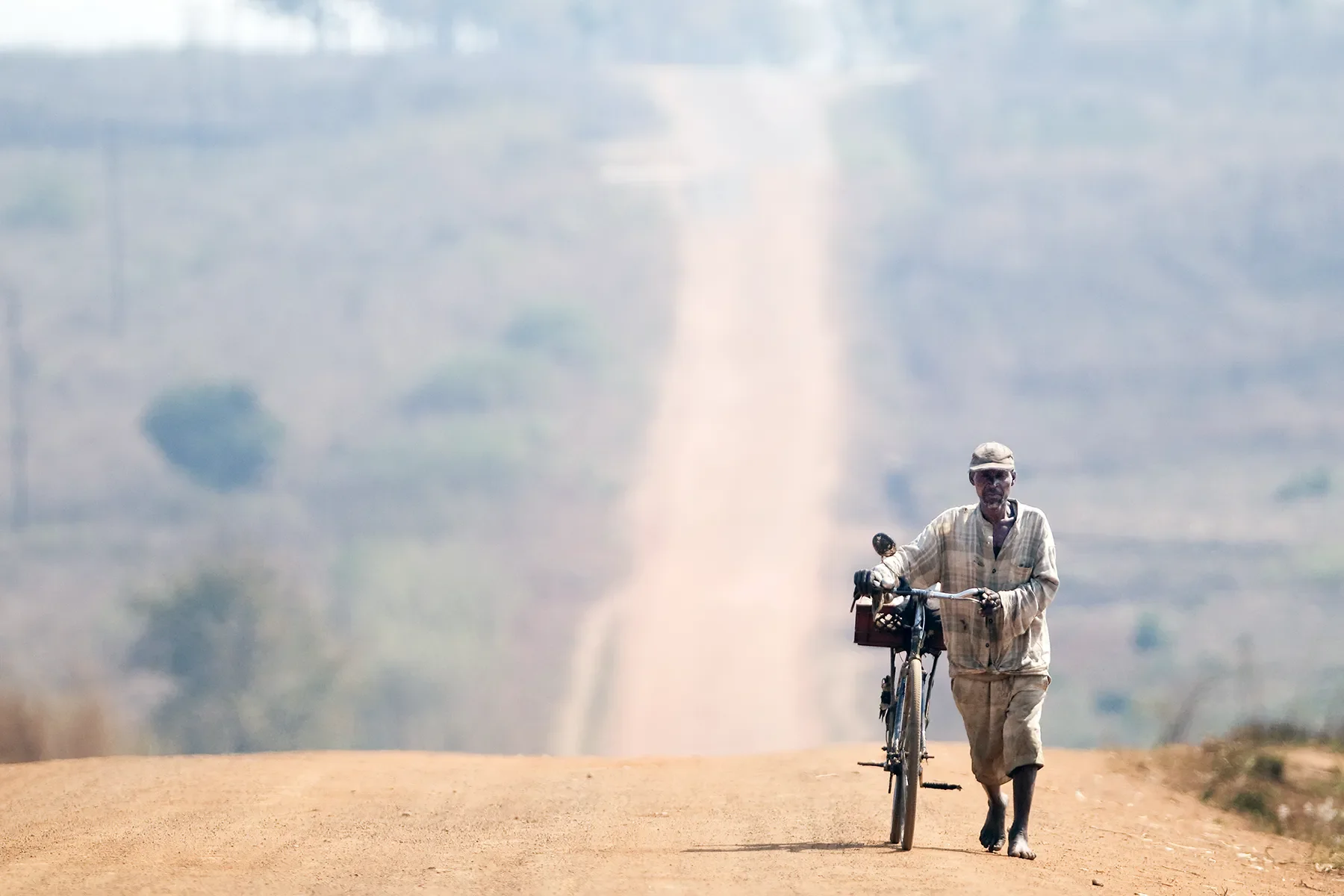 An elderly man walking barefoot on a dirt road, pushing a bicycle.