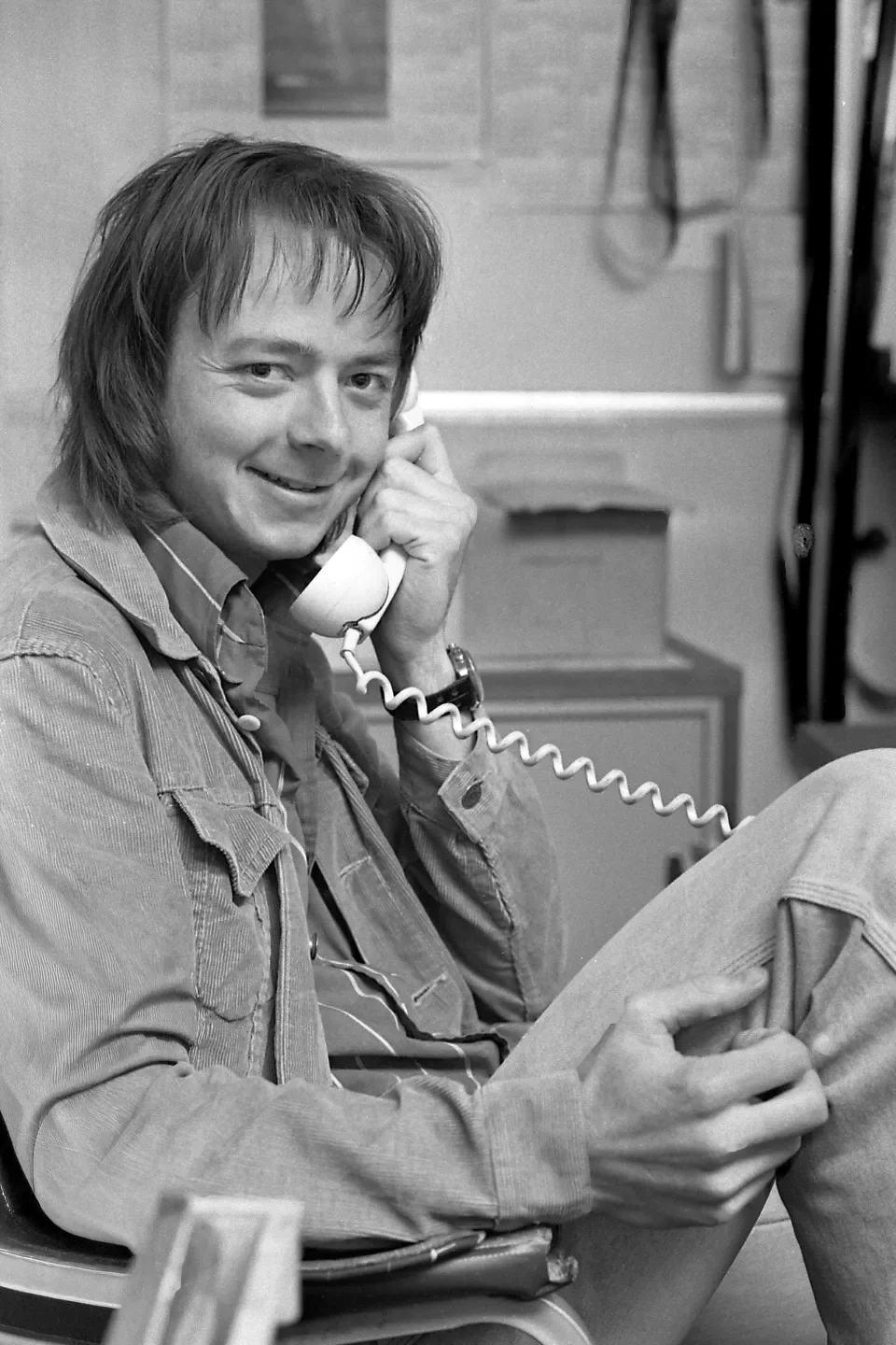 A man with shoulder-length hair smiling while talking on a landline phone, sitting in a chair in what appears to be a casual indoor setting.
