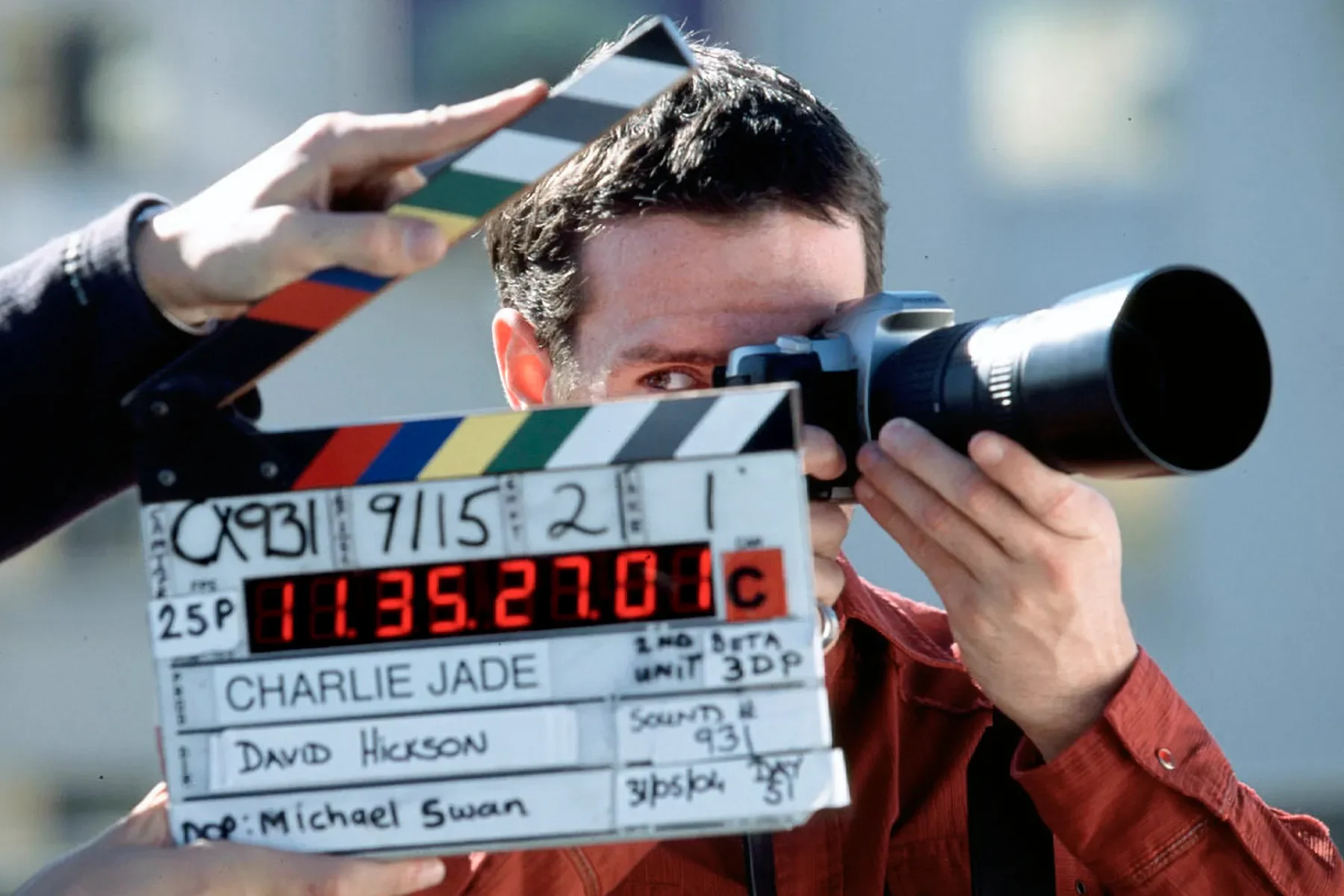 A filmmaker is looking through a camera with a viewfinder, holding a clapperboard with red digital time code and handwritten details in front of him. The background is blurred outdoors.