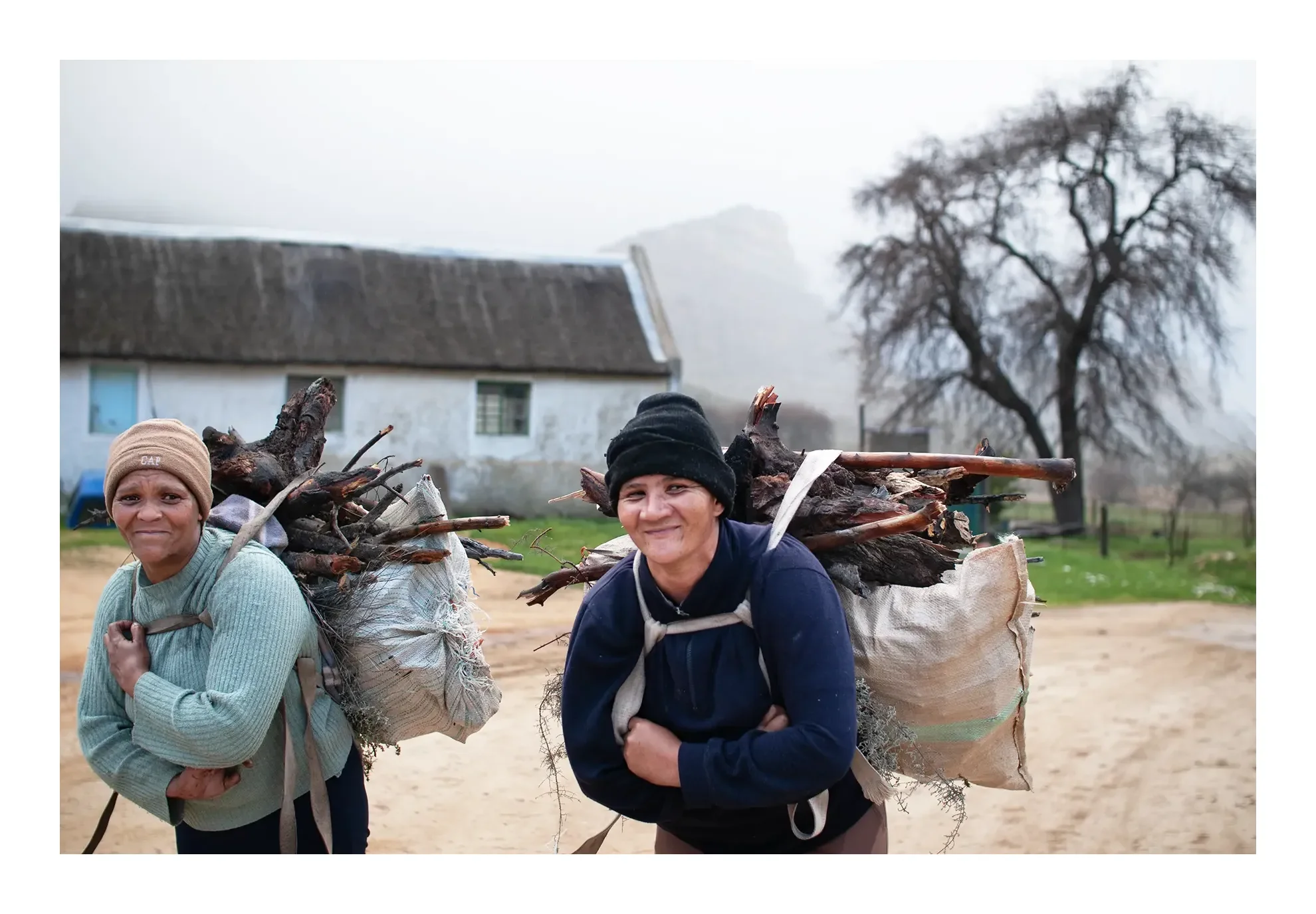 Two women carrying large bundles of firewood on their shoulders outdoors on a cloudy day.