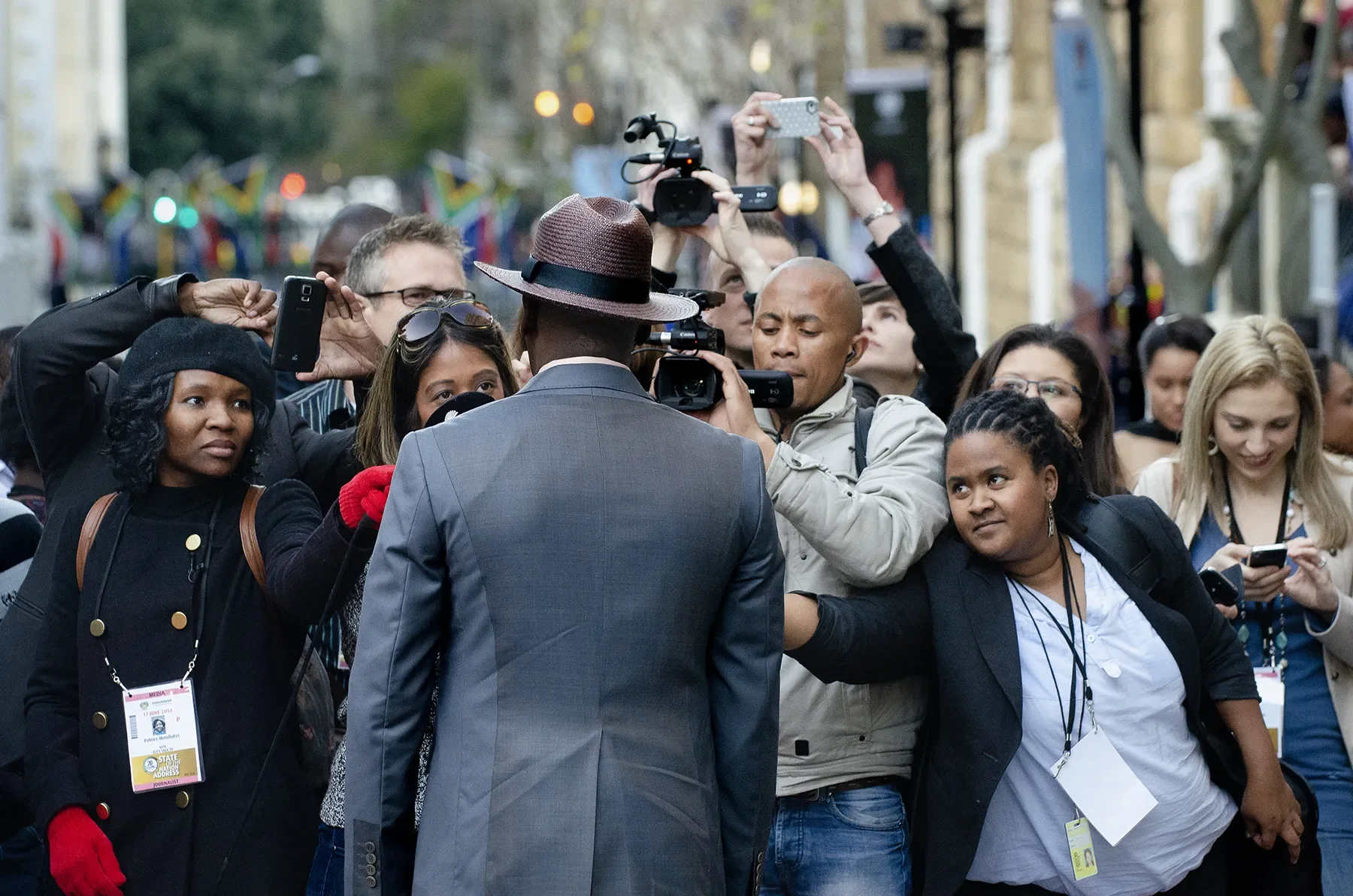 A group of reporters and photographers surrounding and interviewing a man in a grey suit and fedora hat on a city street.