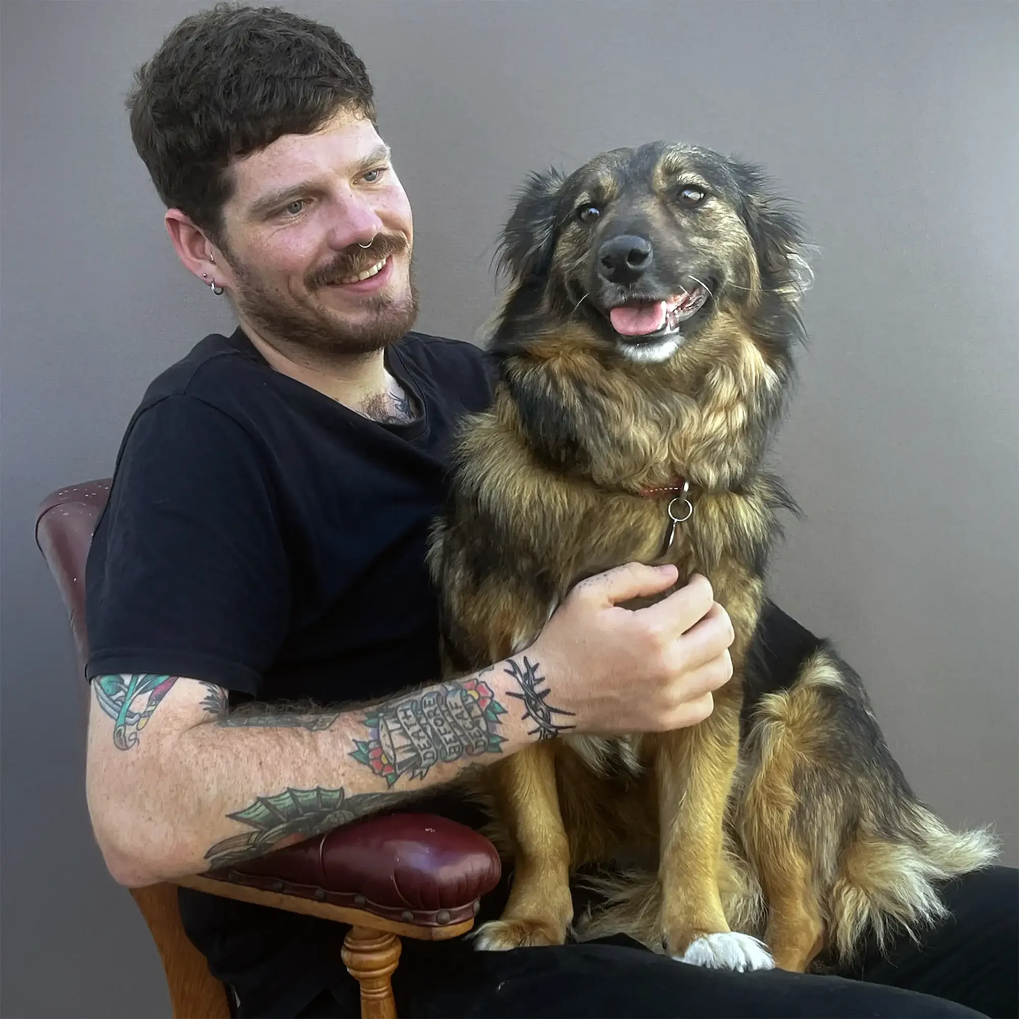 A man with tattoos on his arms sitting on a chair and holding a happy dog on his lap, against a plain gray background.