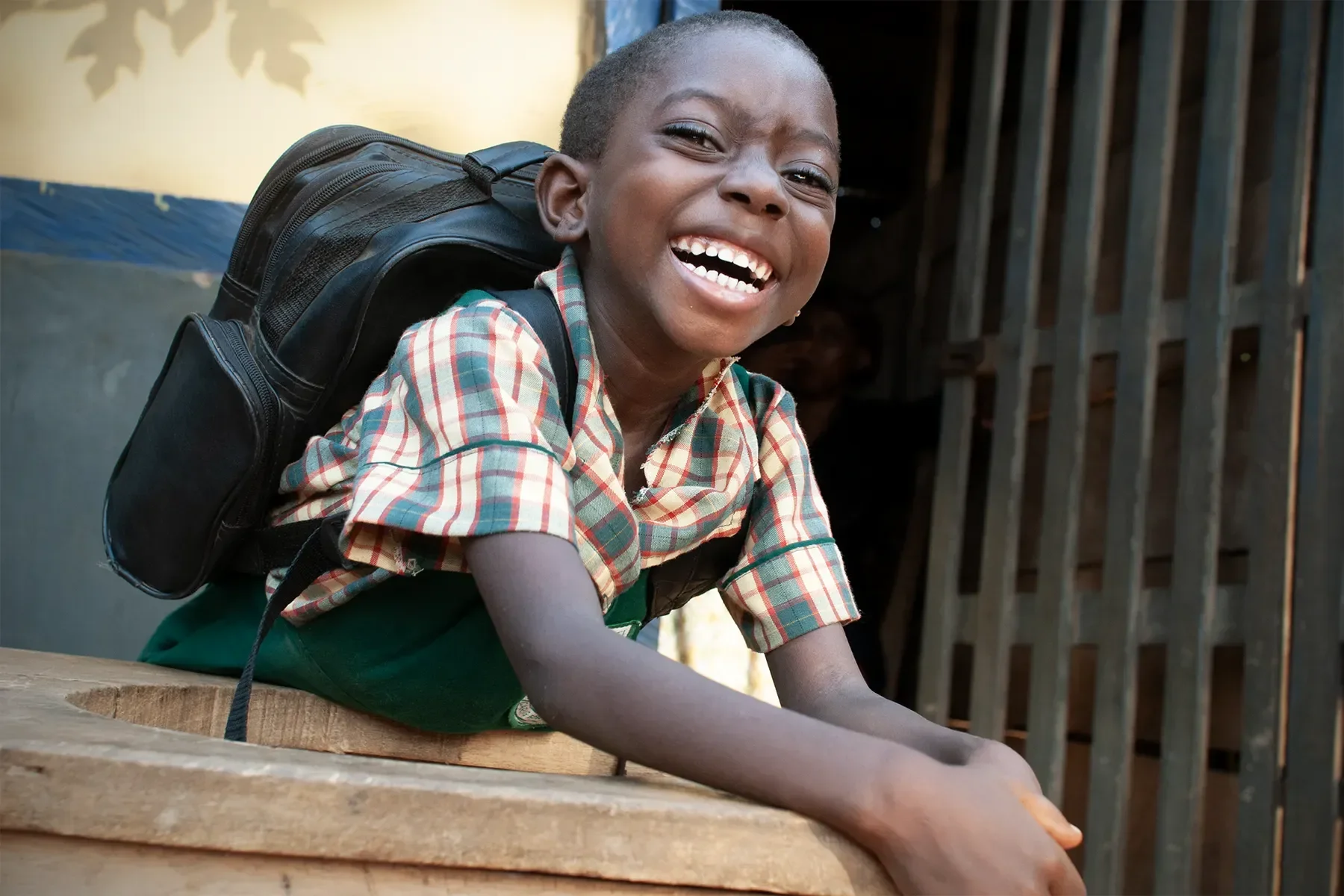A young boy in a plaid shirt with a backpack crouches on a wooden surface, smiling brightly.