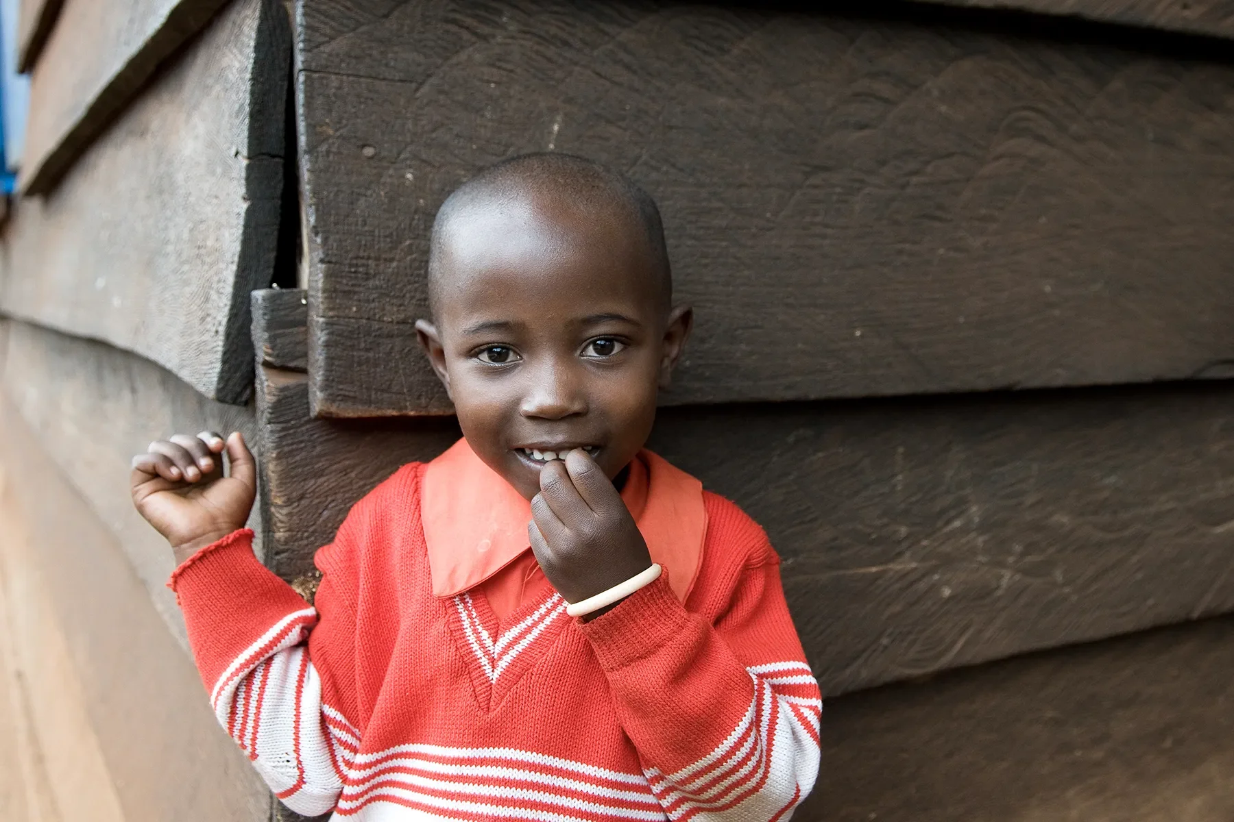 A young boy with a shaved head, wearing a red and white sweater, standing against a wooden wall, smiling and touching his lips.