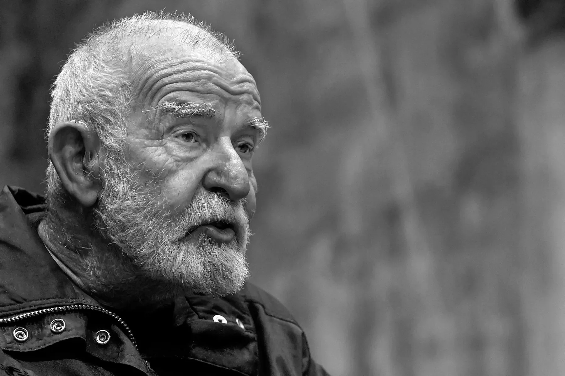 Close-up black and white photo of an elderly man with a beard, short hair, visible forehead wrinkles, and wearing a jacket, looking to the left.