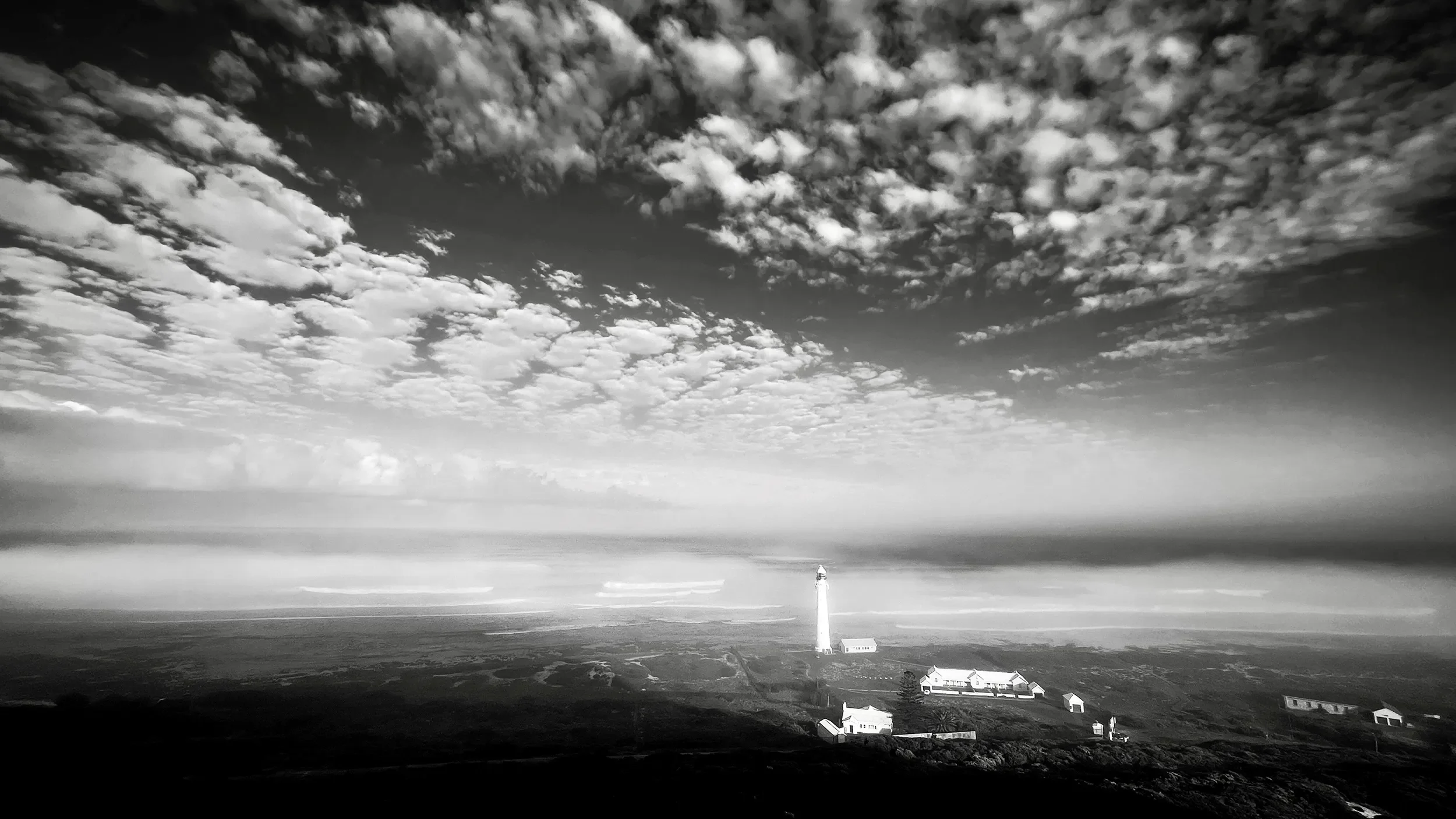 A black and white photo of a lighthouse on a cloudy sky, with some buildings and a landscape in the foreground.