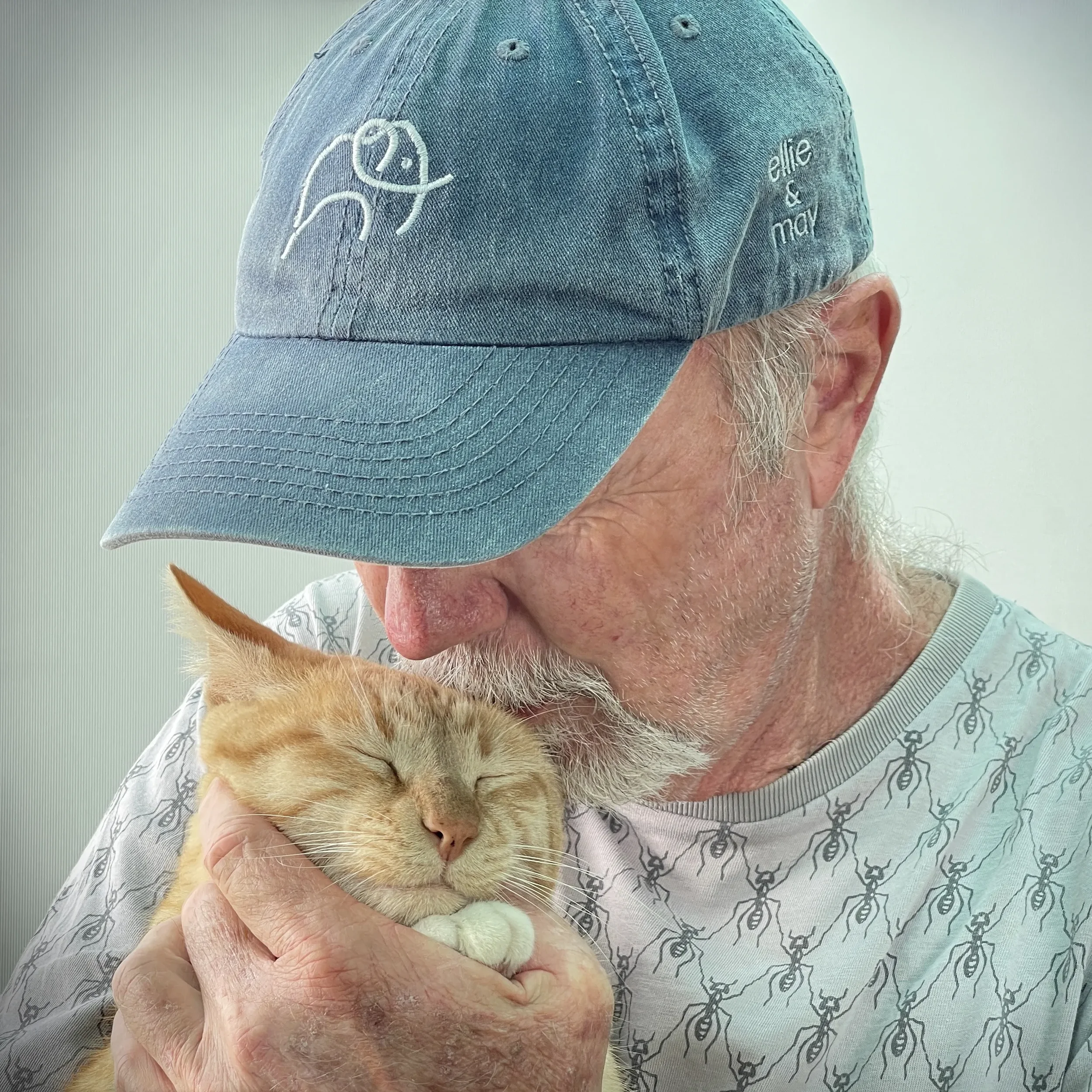 An elderly man with a white beard wearing a blue baseball cap and a white t-shirt holding a sleeping orange tabby cat close to his face, both appear content.