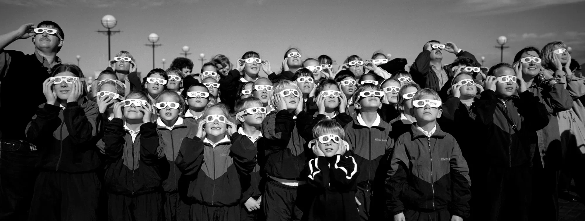 A group of children and adults wearing eclipse glasses, looking up at the sky during a solar eclipse.