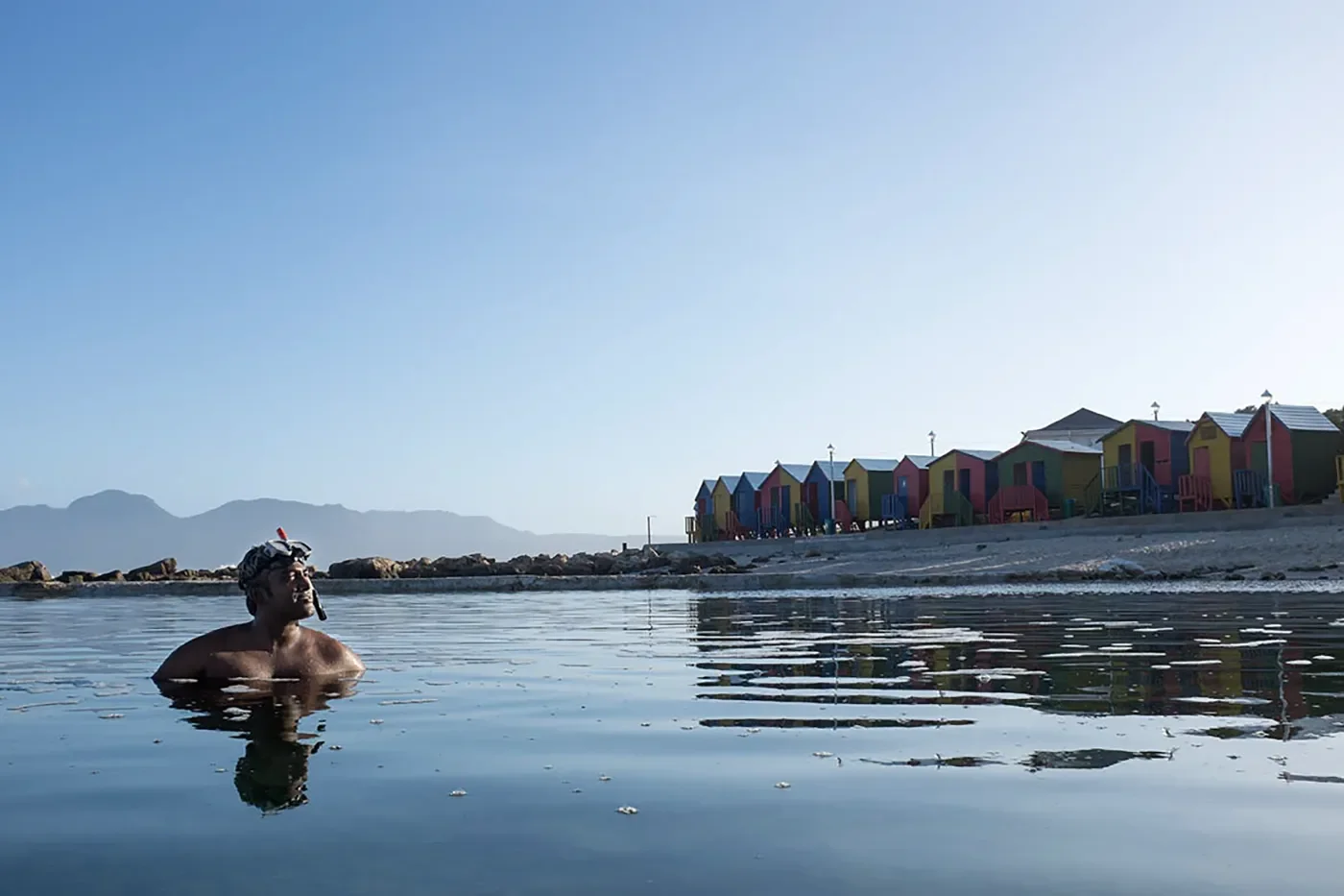 A man in swimwear and a swim cap relaxed in the water near a beach with colorful beach huts and mountains in the background