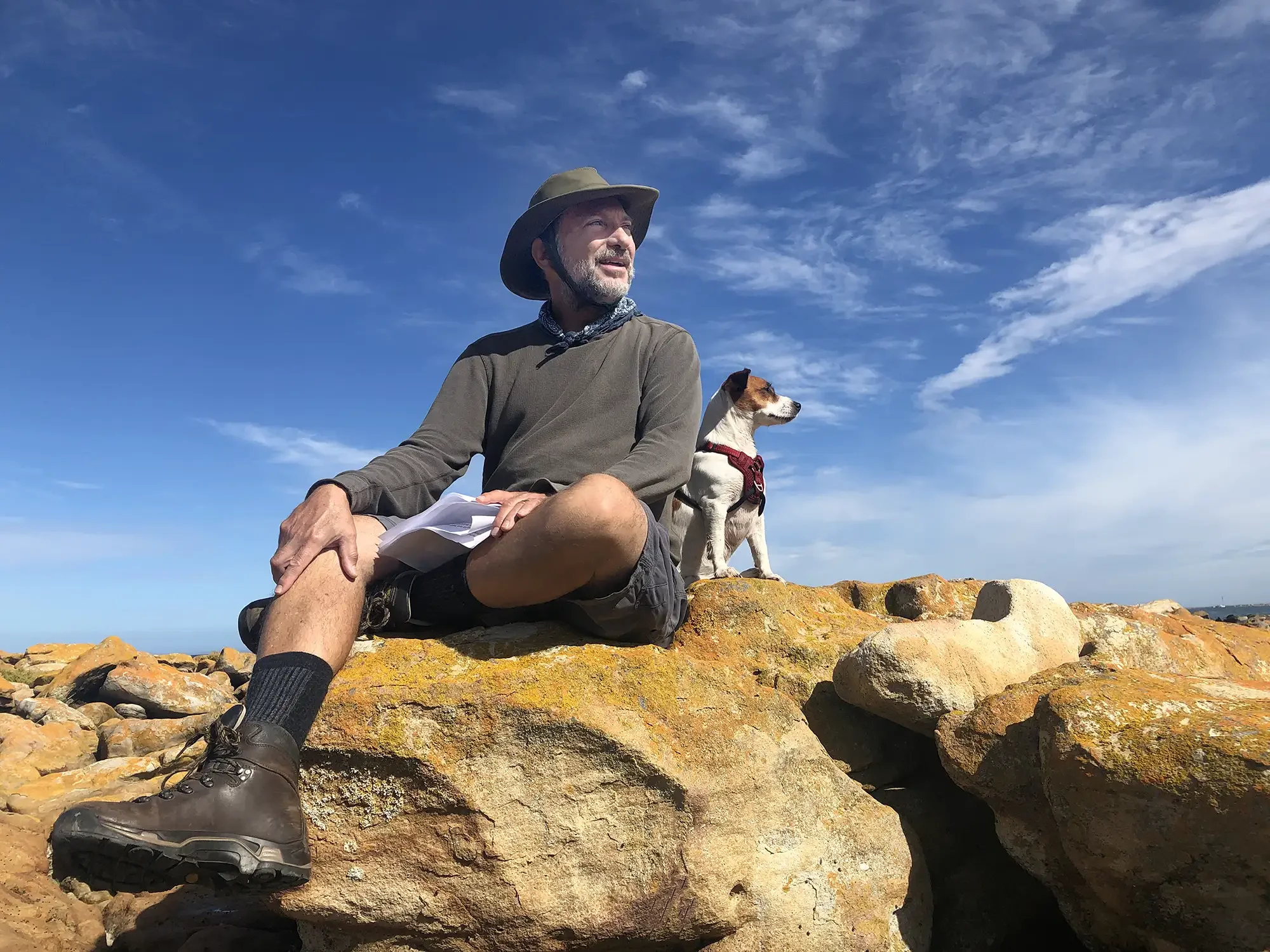 A man and a dog sitting on large rocks outdoors under a blue sky with scattered clouds. The man is wearing a wide-brimmed hat, long-sleeved shirt, shorts, and hiking boots. The dog, wearing a red harness, is seated next to the man, both looking into 