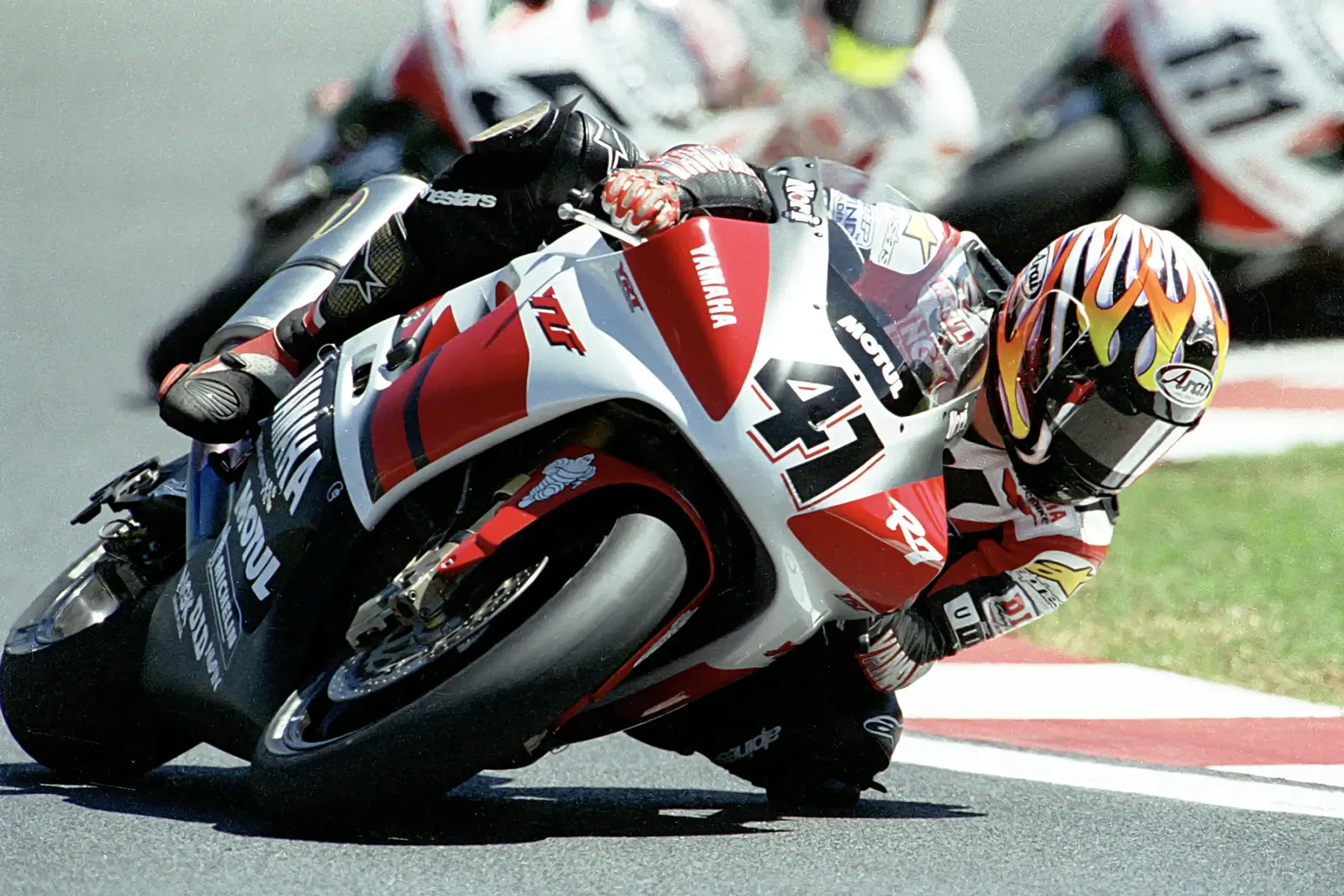 Motorcycle racer leaning into a turn on a racetrack, wearing a colorful helmet and racing suit, with other racers in the background.