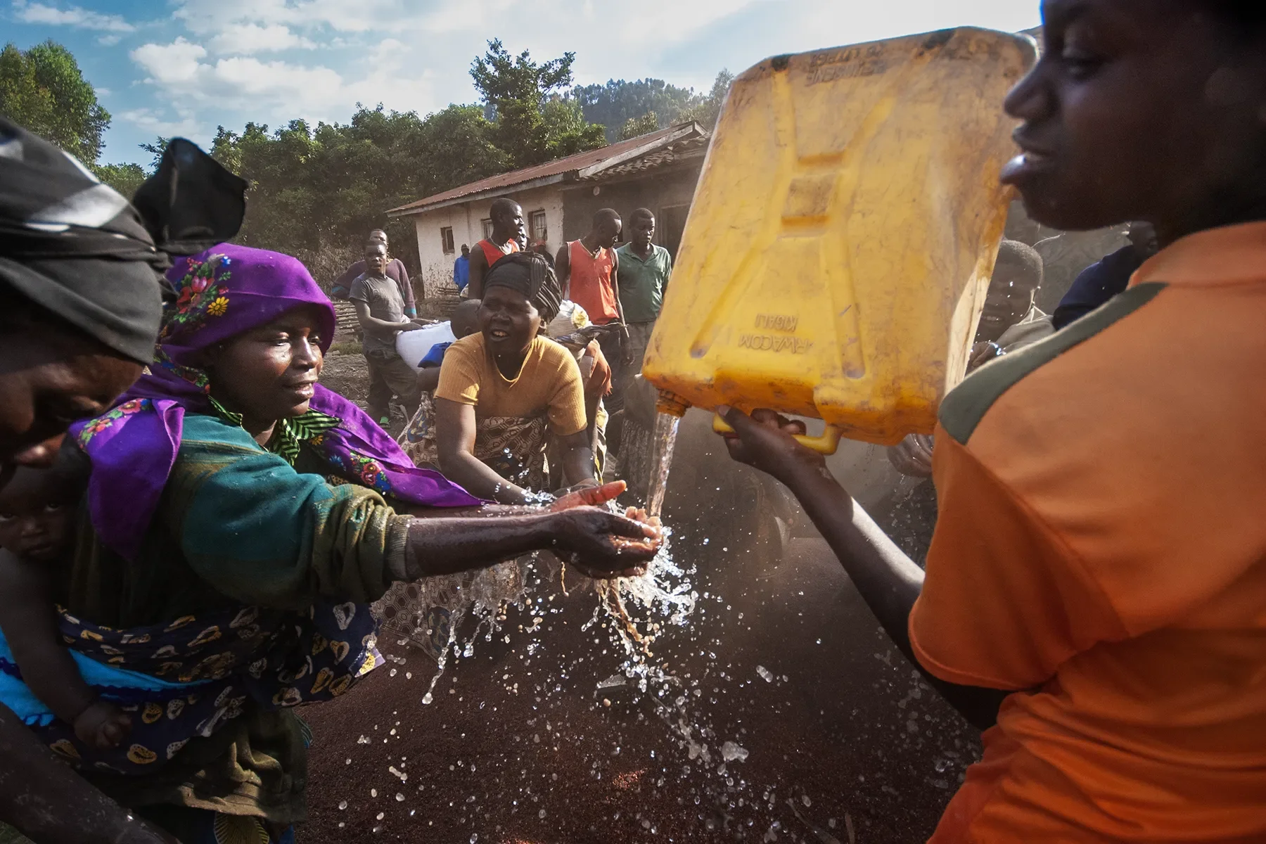 A group of people in an outdoor setting, with some taking water from a yellow container poured by a person in an orange shirt. Others are observing or waiting nearby, and a house with a metal roof is visible in the background.