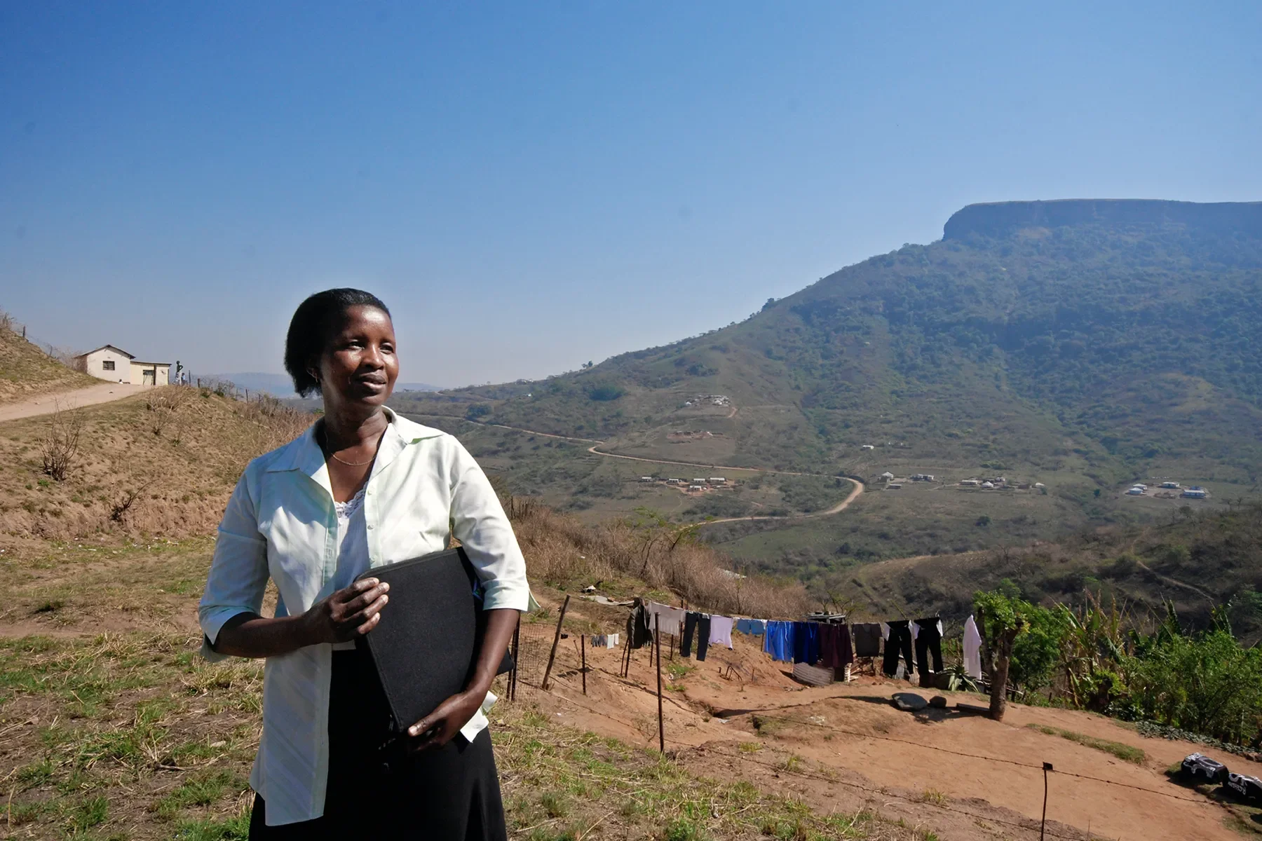 A woman standing outdoors in a rural area with hills in the background. She is holding a black folder and looking to her left. There are laundry clothes hanging on a line nearby, and small houses and a winding dirt road can be seen on the hillside.