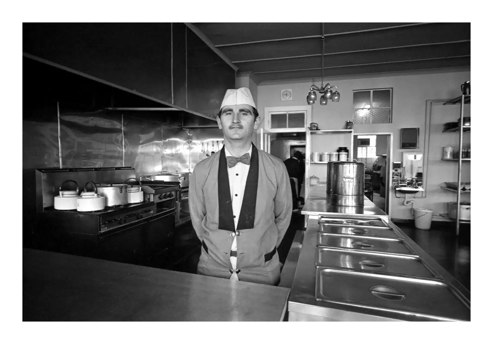 Black and white photo of a man in a vintage restaurant kitchen, wearing a chef's hat, bow tie, and apron, standing behind a counter with kitchen equipment and shelves in the background.