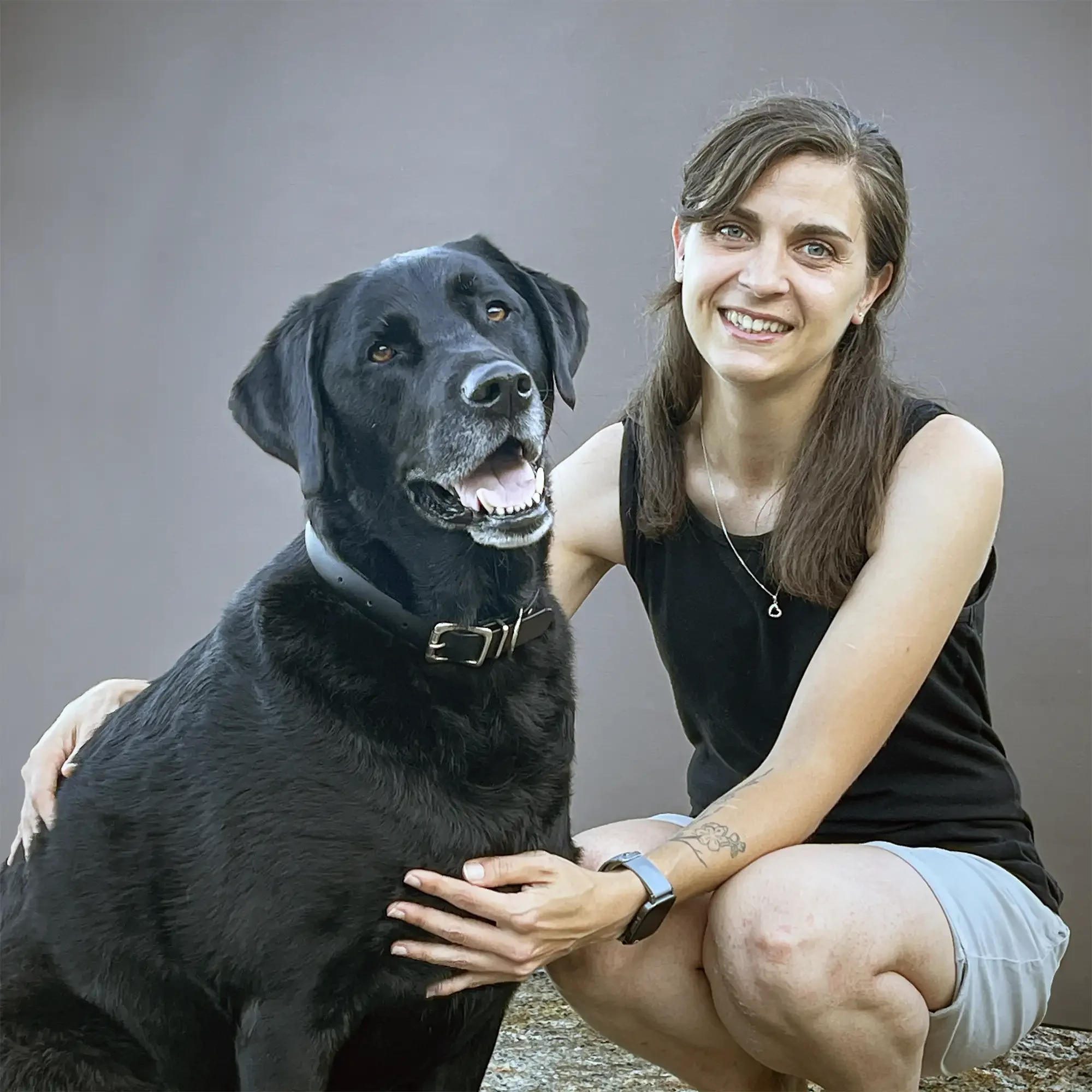 A woman smiling and sitting next to a black Labrador retriever dog with a gray collar.