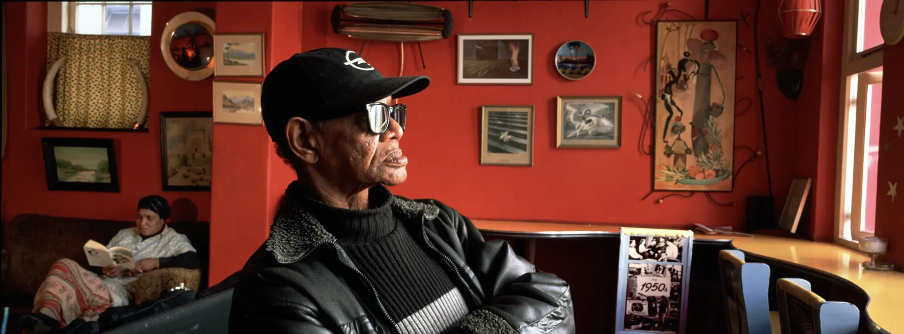An elderly man wearing a black cap, sunglasses, and a black leather jacket sitting in a colorful room with red walls. In the background, a woman is sitting on a couch reading a book, with various framed pictures and artwork hanging on the wall.