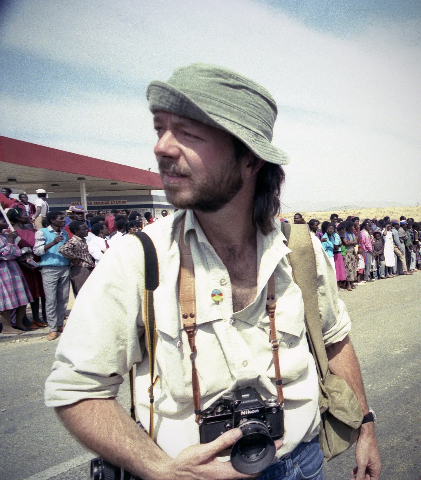 A man with a Nikon camera hanging from his neck, wearing a light-colored shirt, a bucket hat, and carrying a backpack, standing in front of a crowd of people at a busy outdoor location with a service station in the background.