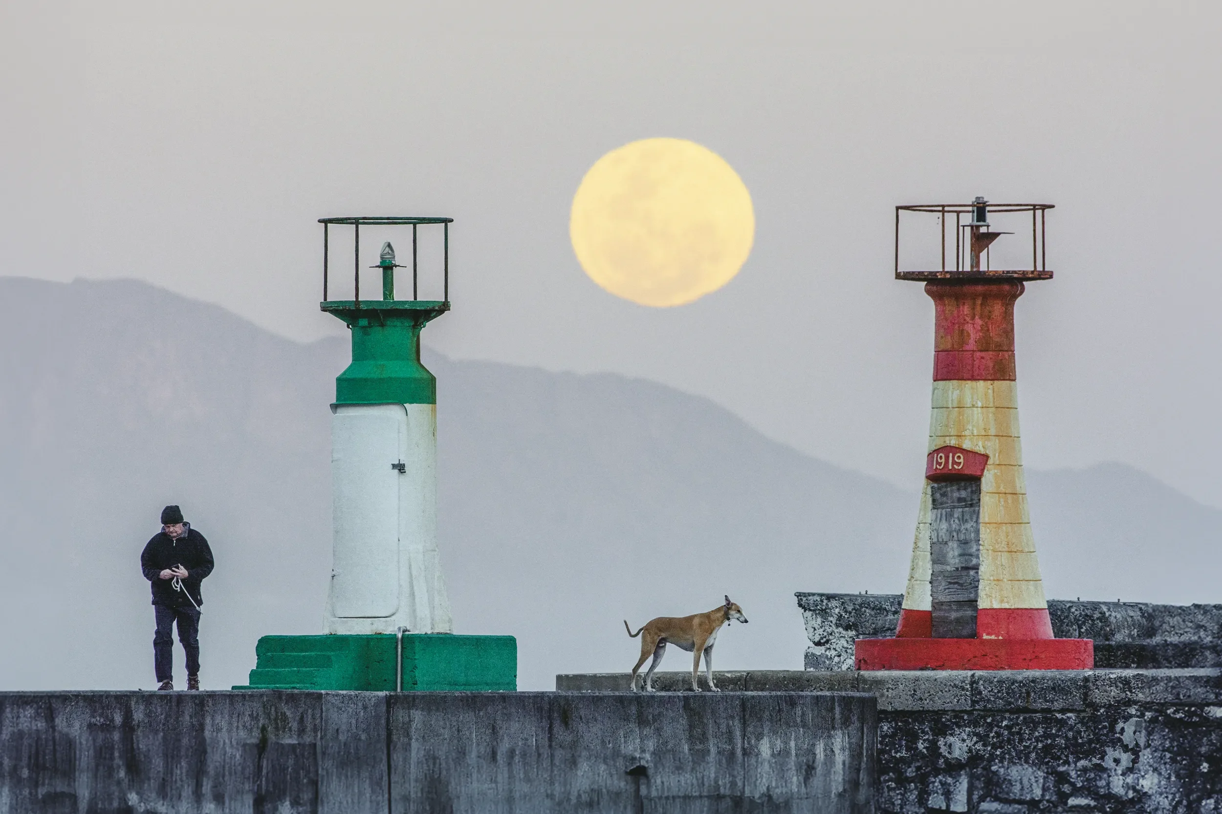 A person walks a dog on a concrete pier with two colorful lighthouses and a large full moon in the sky behind them.