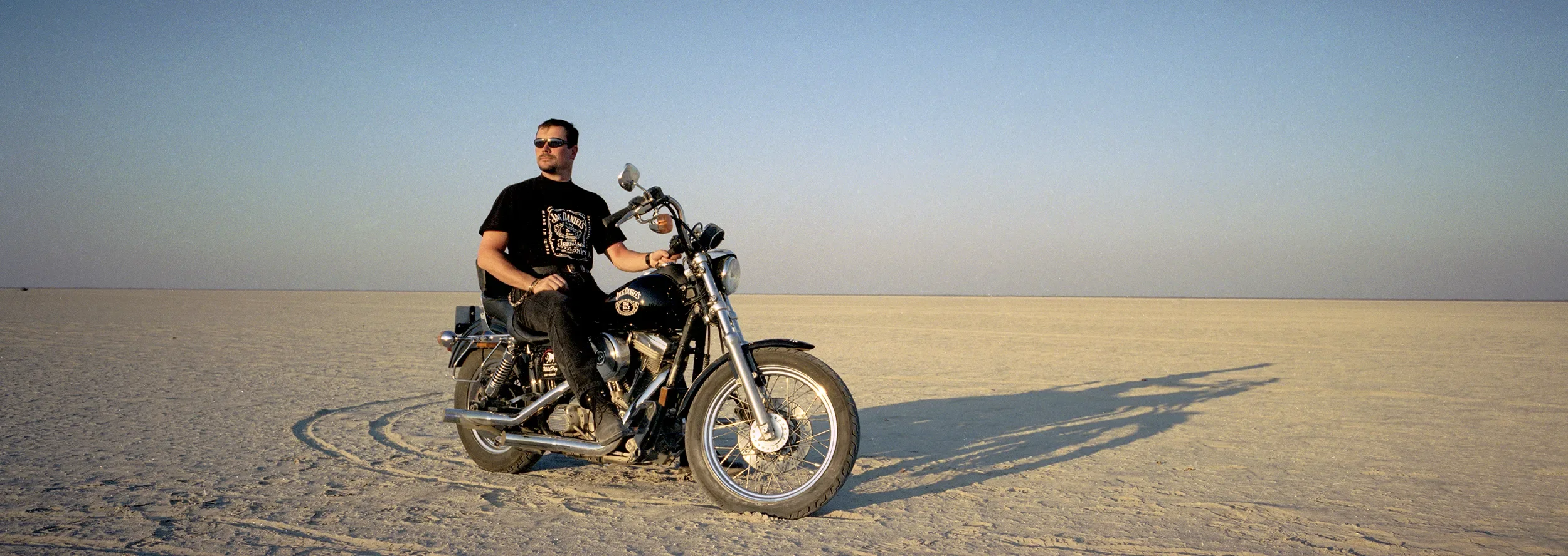 Man sitting on a black Harley-Davidson motorcycle in a vast, flat desert landscape with a clear sky.