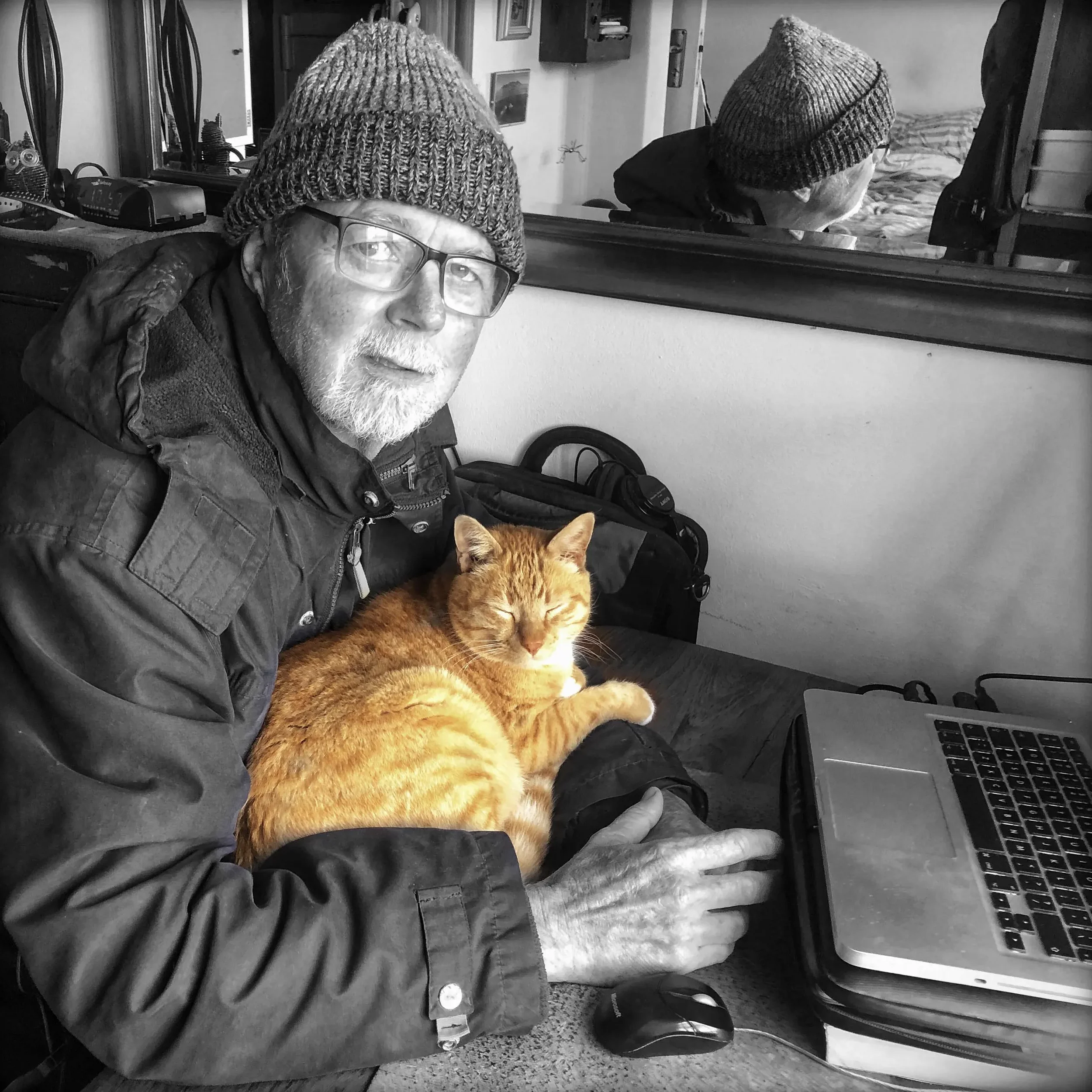 An elderly man wearing a beanie, glasses, and a jacket, sitting at a desk with a ginger cat resting on his lap, in a room with a mirror and various objects in the background, all in black and white except the cat in color.
