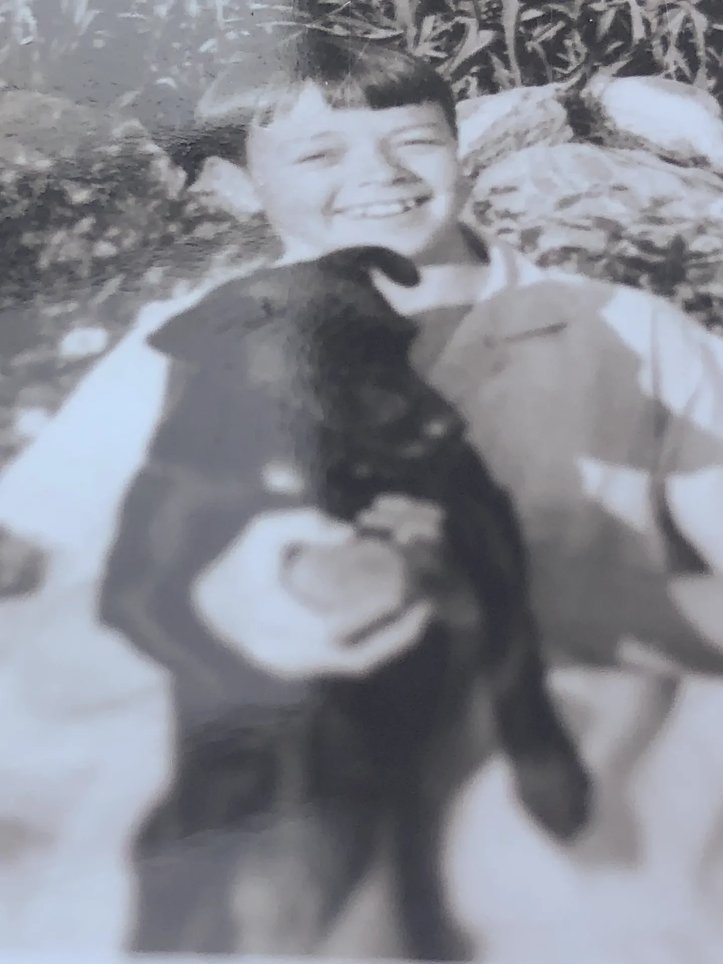 A black-and-white photograph of a young boy with short hair, smiling broadly, holding a small dog in his arms. The background features rocks or stones.