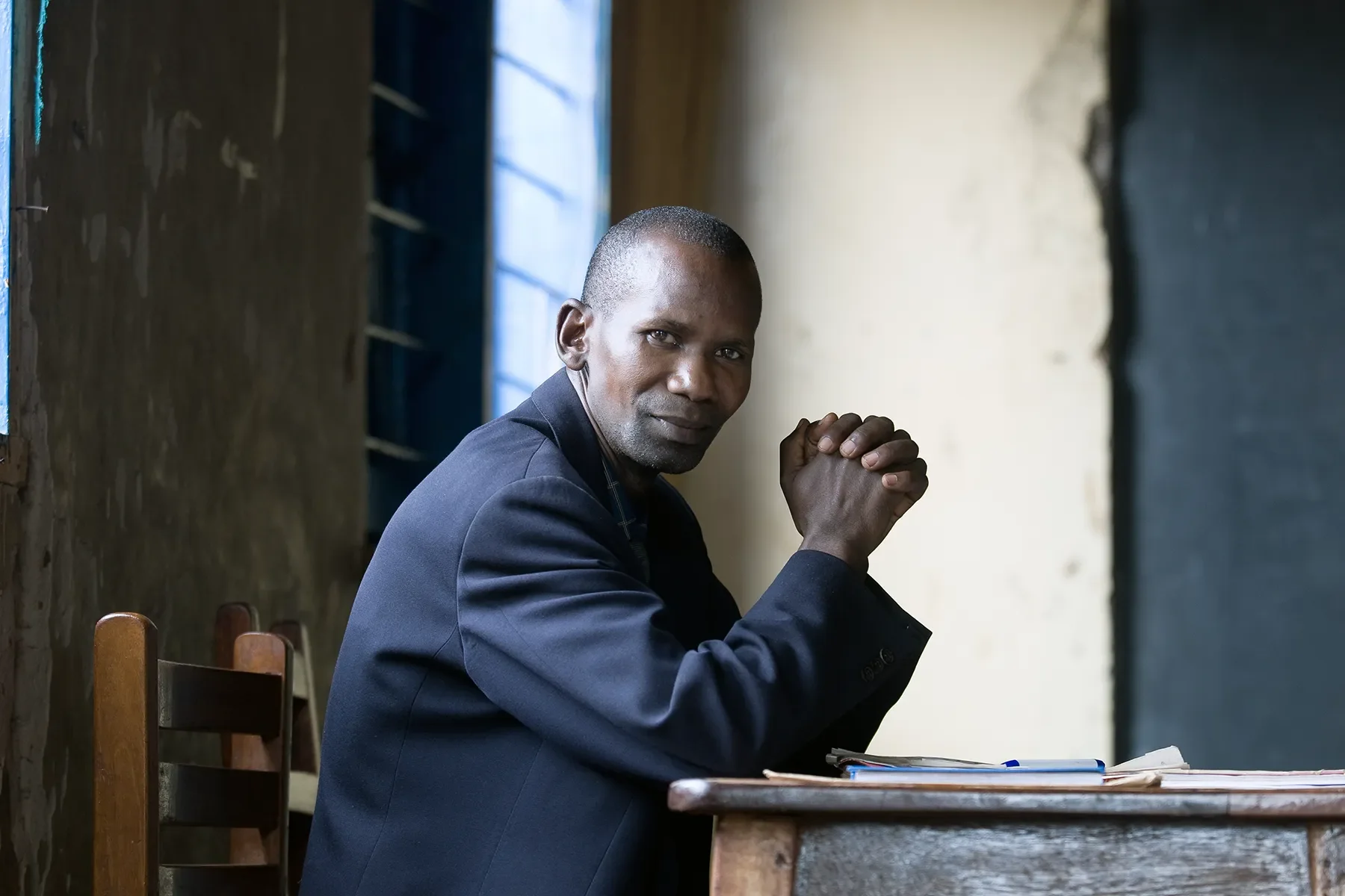 A man in a dark suit sitting at a wooden table with his hands clasped, looking into the camera with a serious expression.