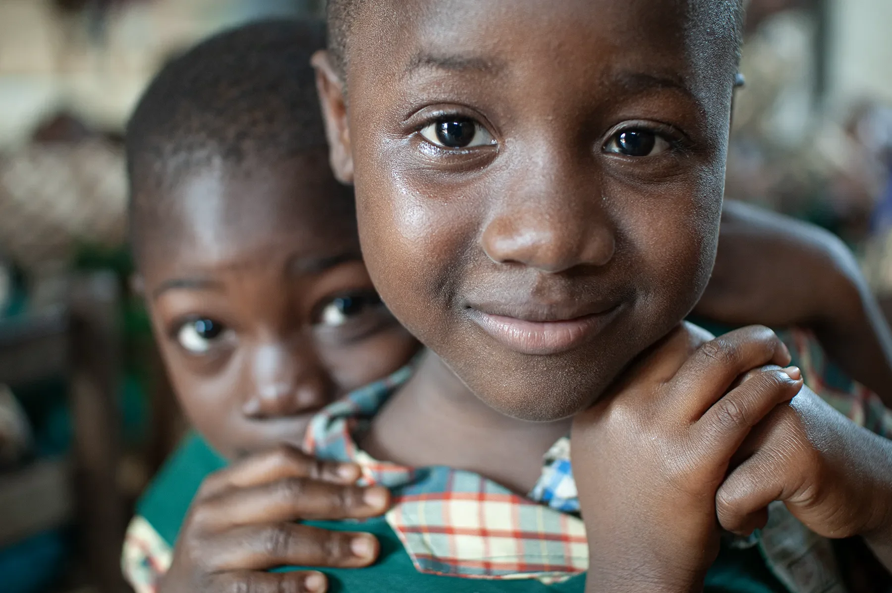 Two children, a girl in the foreground and a boy behind her, smiling and looking at the camera, indoor setting.