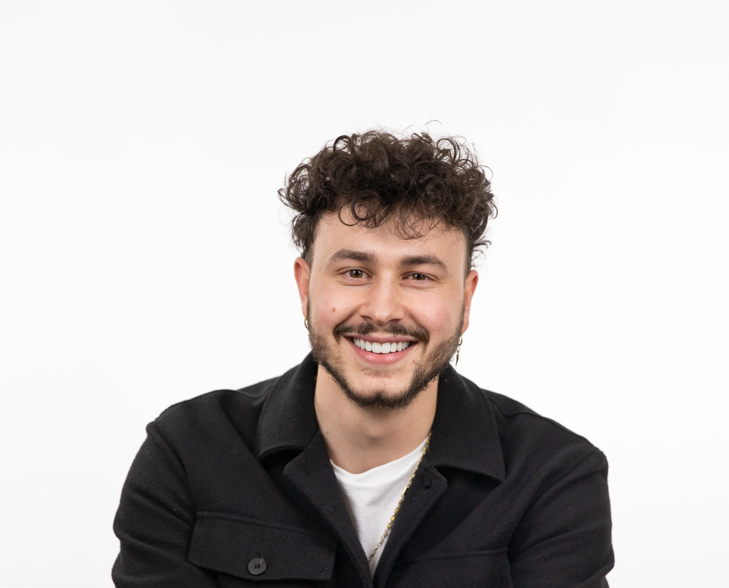 Portrait of a smiling young man with curly dark brown hair, beard, and earrings, wearing a black jacket and a white shirt against a plain white background.