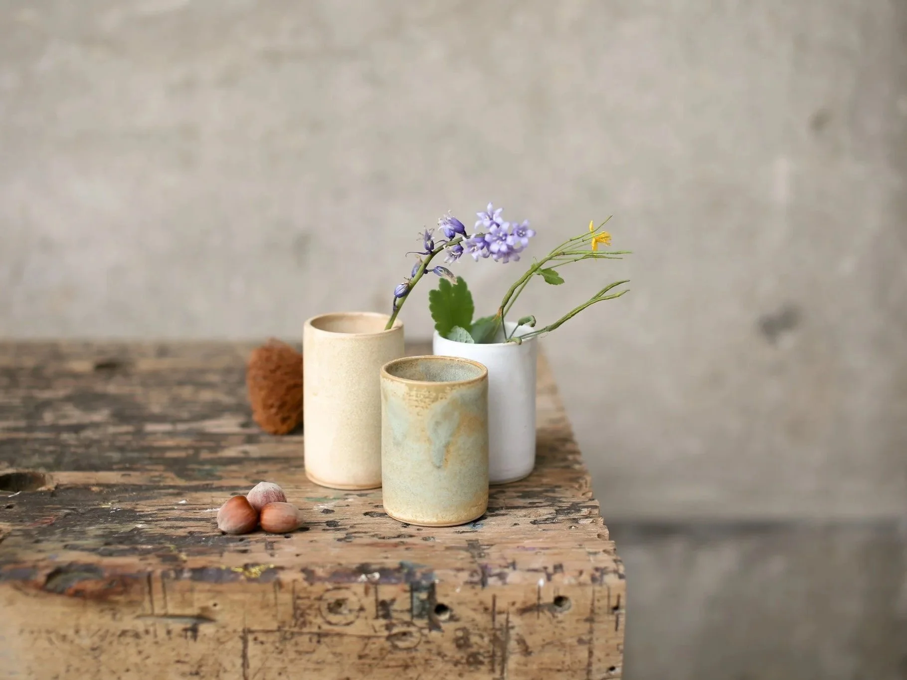Three ceramic vases with flowers and greenery on a weathered wooden table, with hazelnuts in the foreground and a plain wall in the background.
