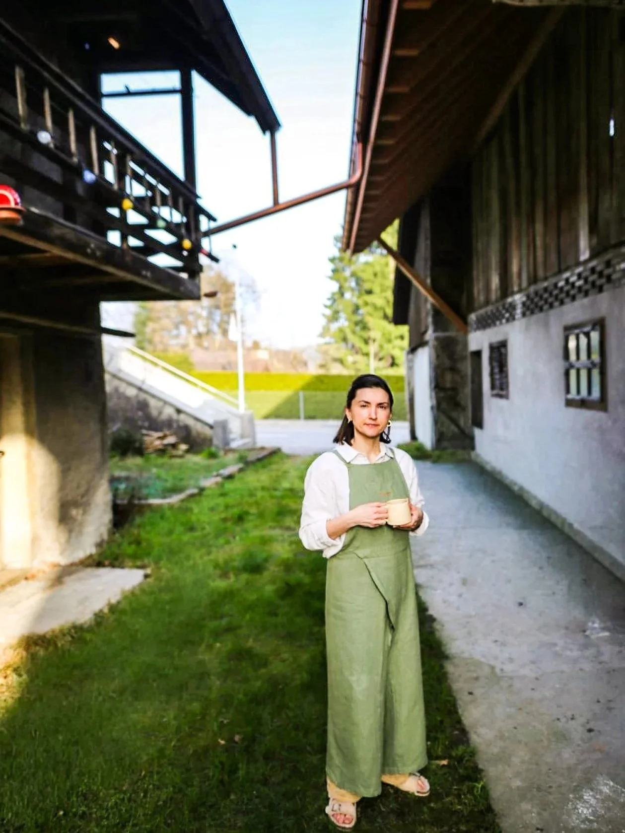 A woman standing outside in a grassy area, holding a cup and looking at the camera. She is wearing a white shirt and green overalls. There are buildings and trees in the background.