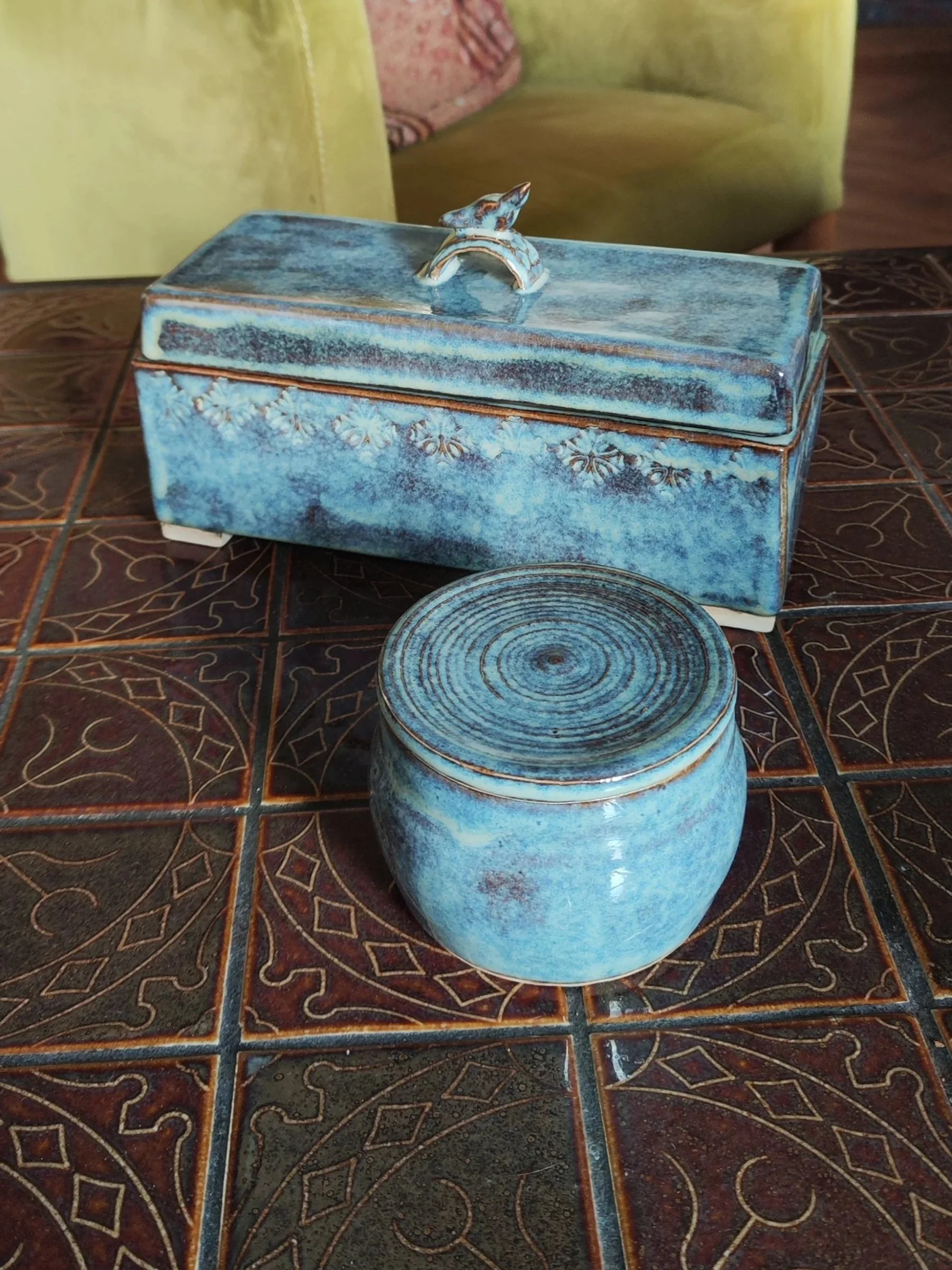 A blue ceramic jewelry box with a bird figurine on its lid, sitting on a patterned tile table.