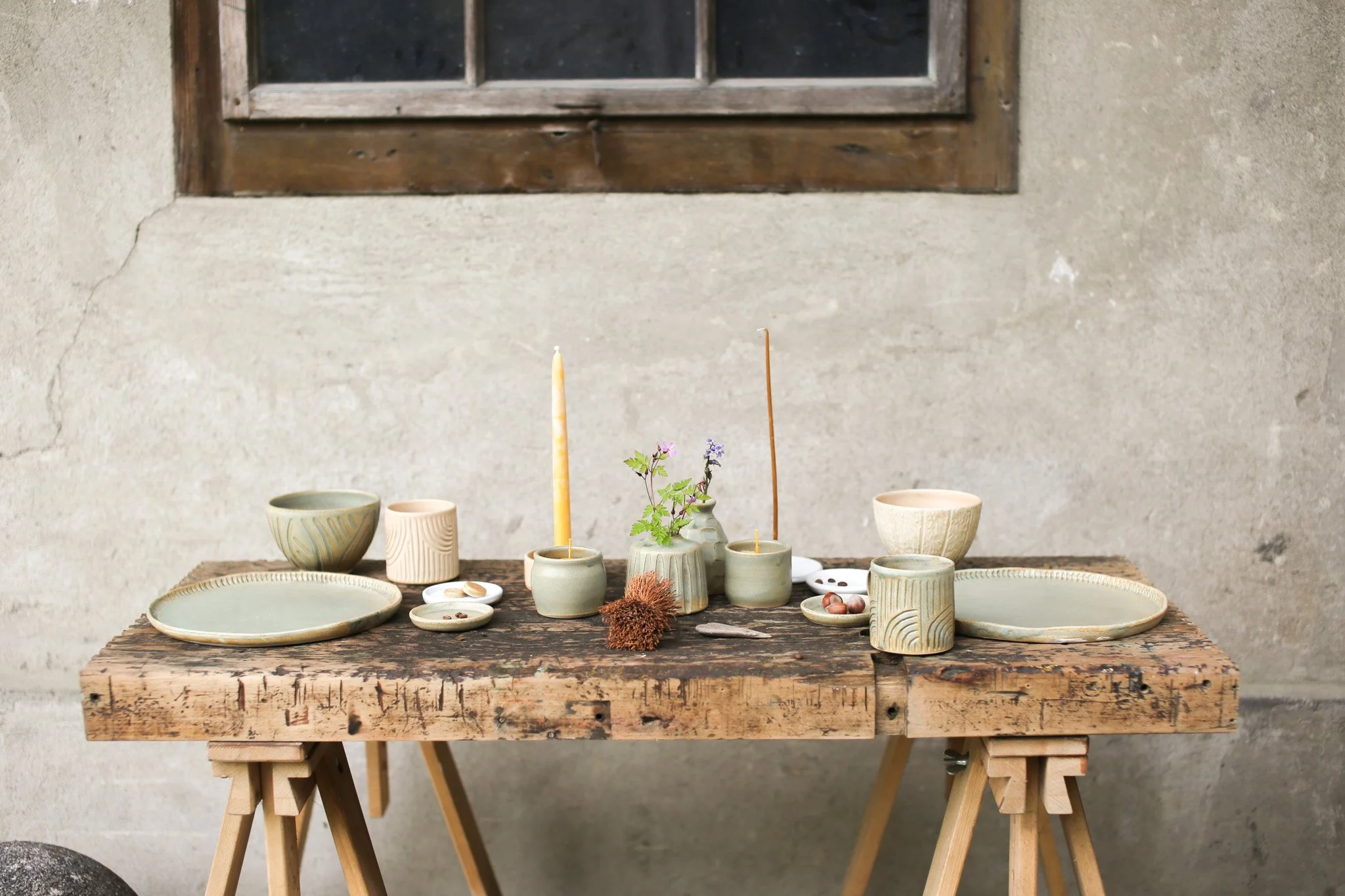 A rustic table displaying ceramic bowls, cups, plates, a small plant, candles, and some seasonal decorations, set against an unfinished concrete wall with a small window.