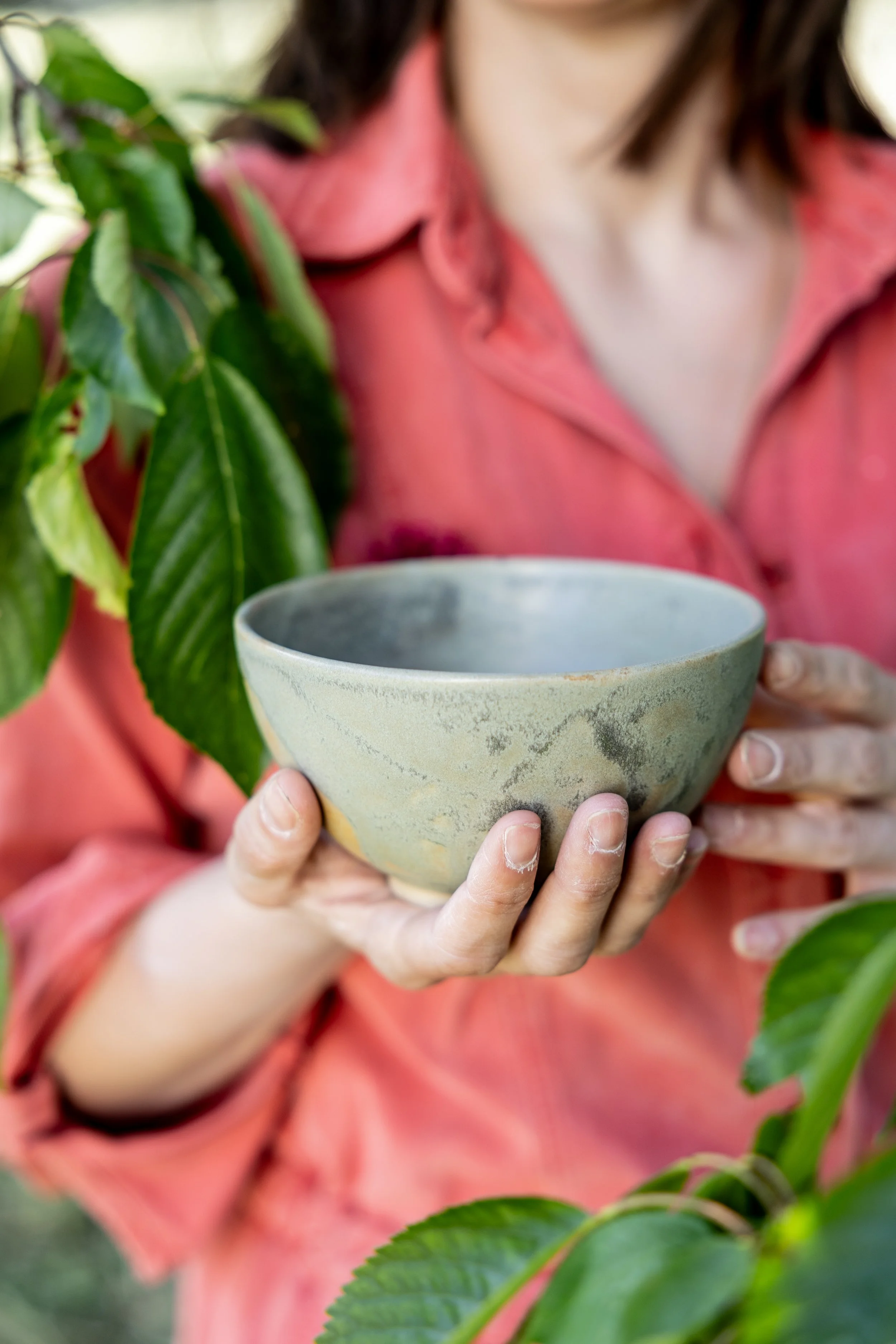 A person holding a ceramic bowl with both hands, surrounded by green leaves. The person is wearing a pink shirt.