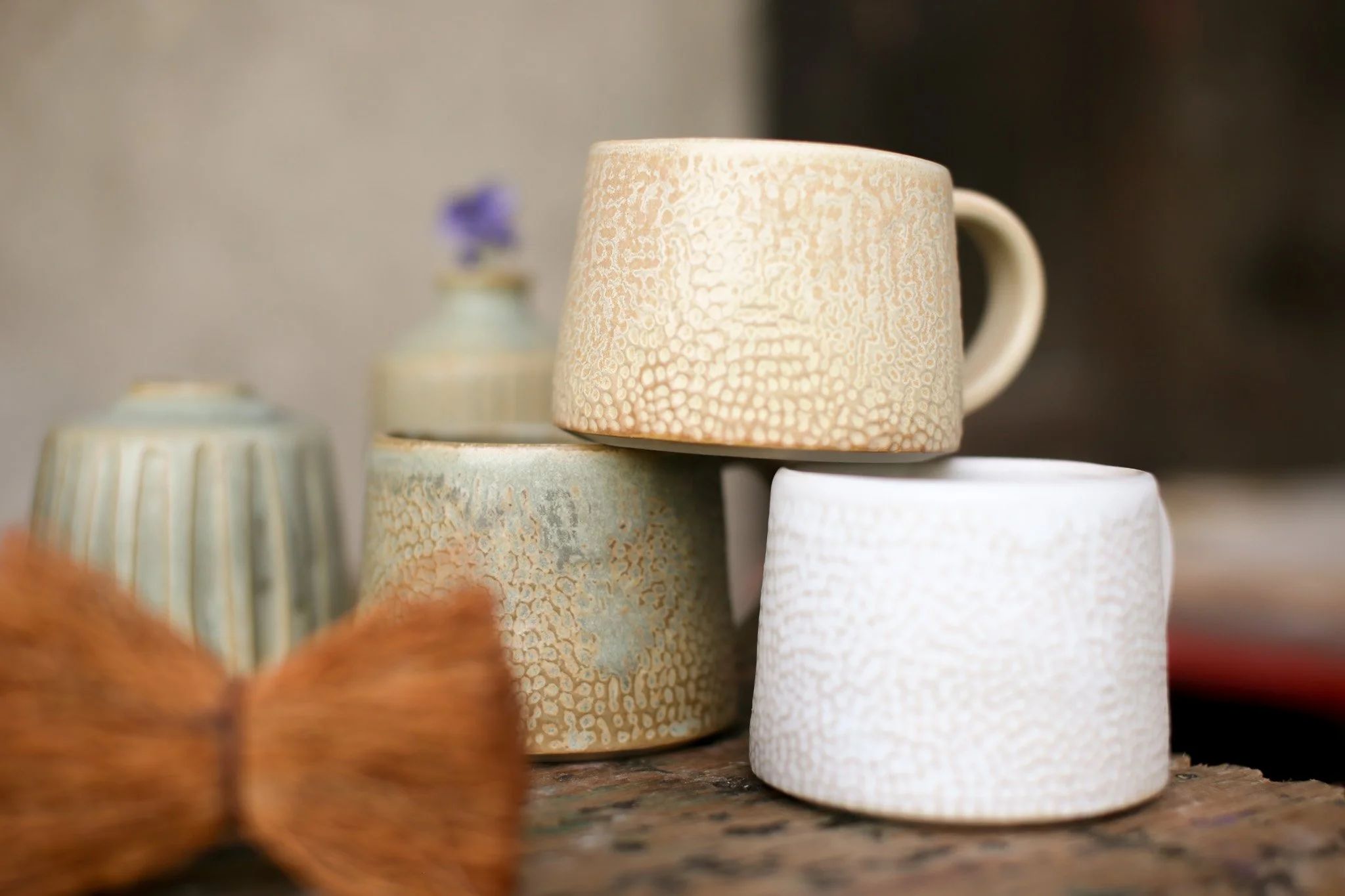 Stacked ceramic cups with textured and glazed surfaces, placed on a wooden surface, with blurred vases and a purple flower in the background.
