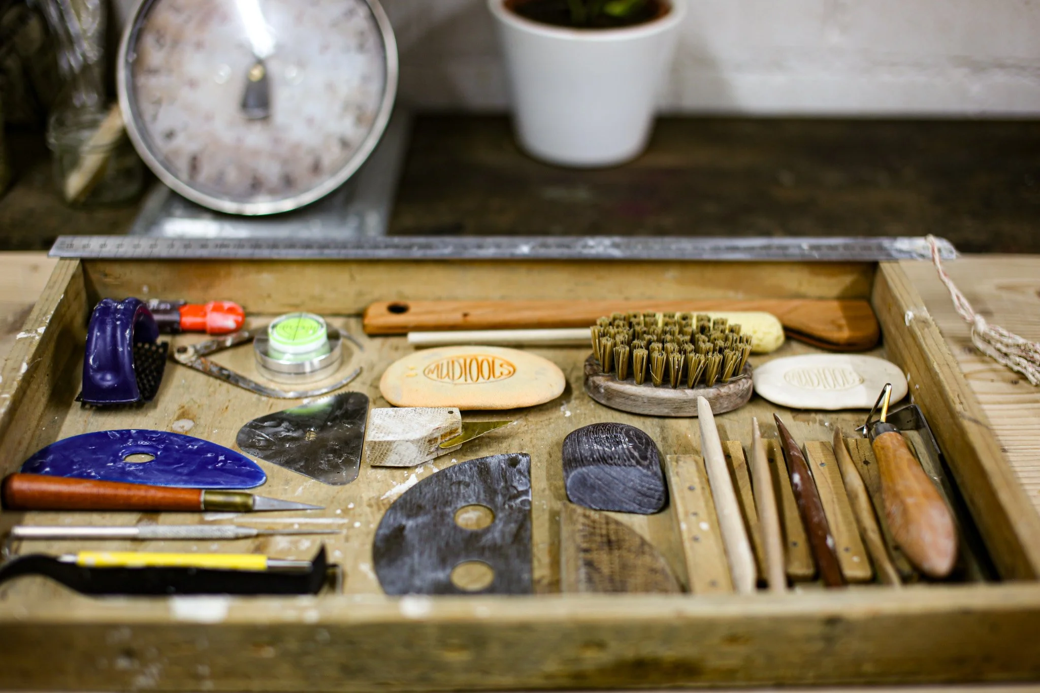 A cluttered wooden workbench with pottery-making tools,  including a blue and a black rib, a brush, and small sponges.