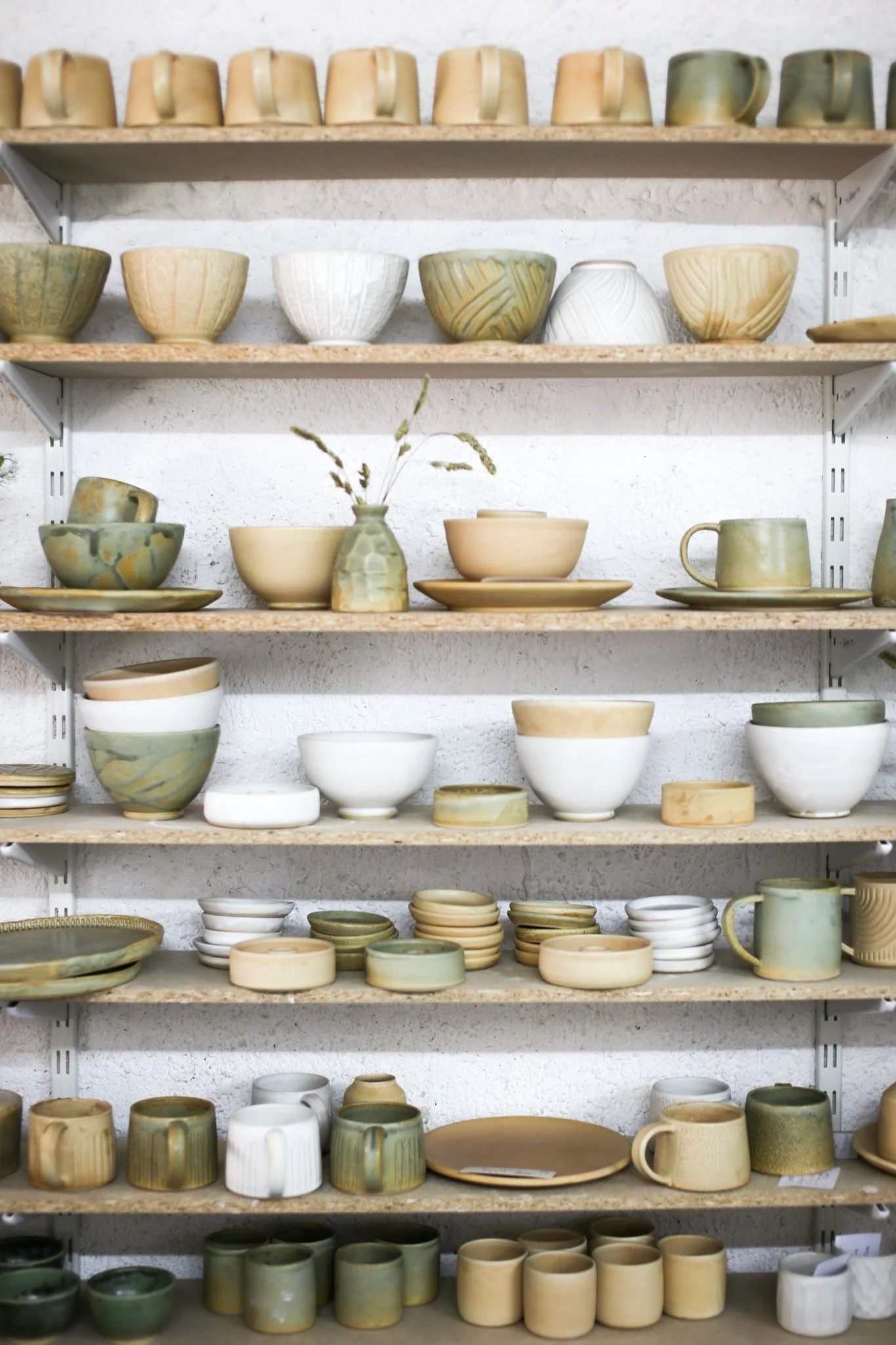Multiple shelves filled with various handmade ceramic cups, bowls, and plates in earth tones, arranged in neat rows against a white wall.