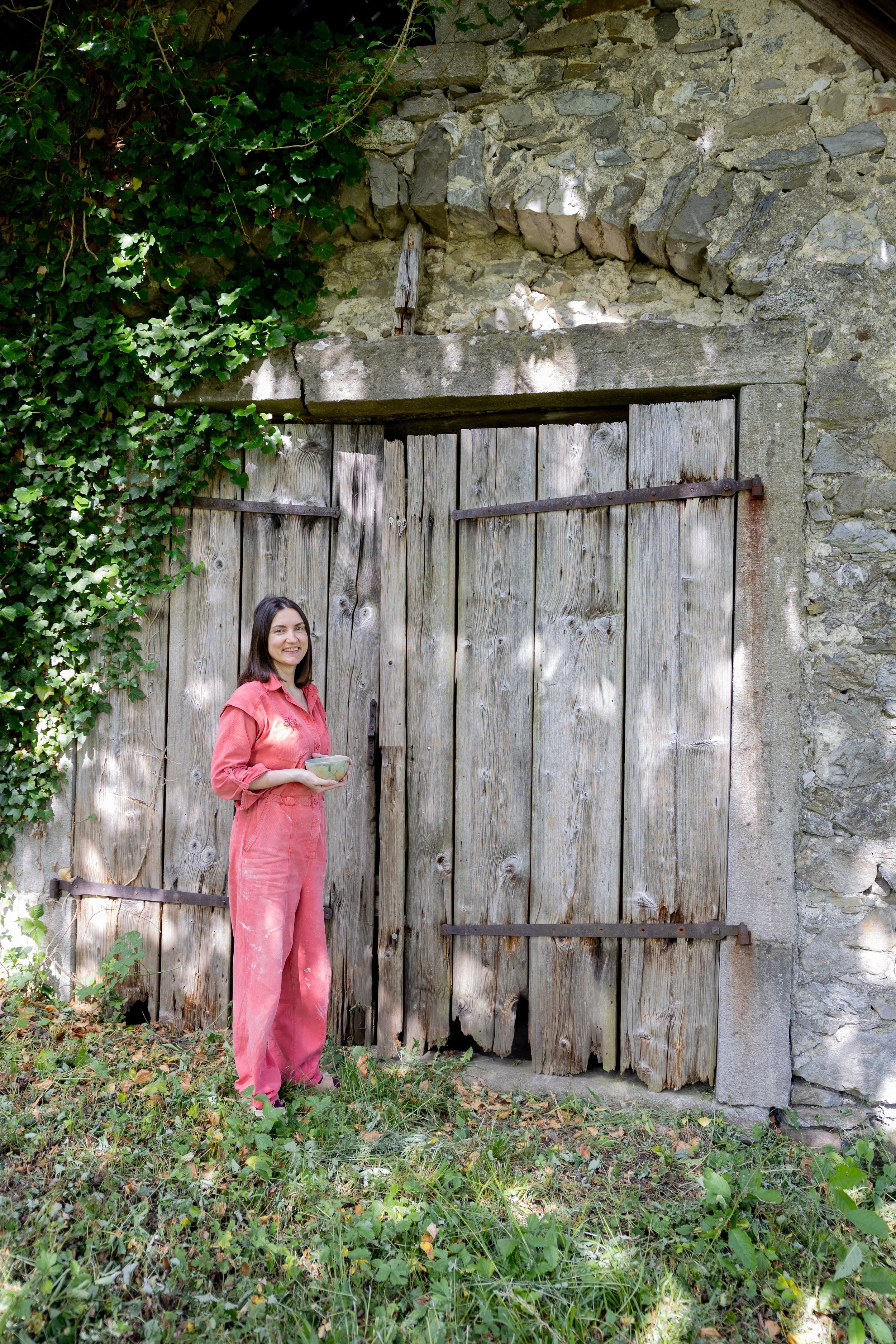 A woman with long dark hair, wearing a pink jumpsuit, standing outside next to a large wooden gate, holding a bowl in her hands, with green foliage and a stone wall in the background.