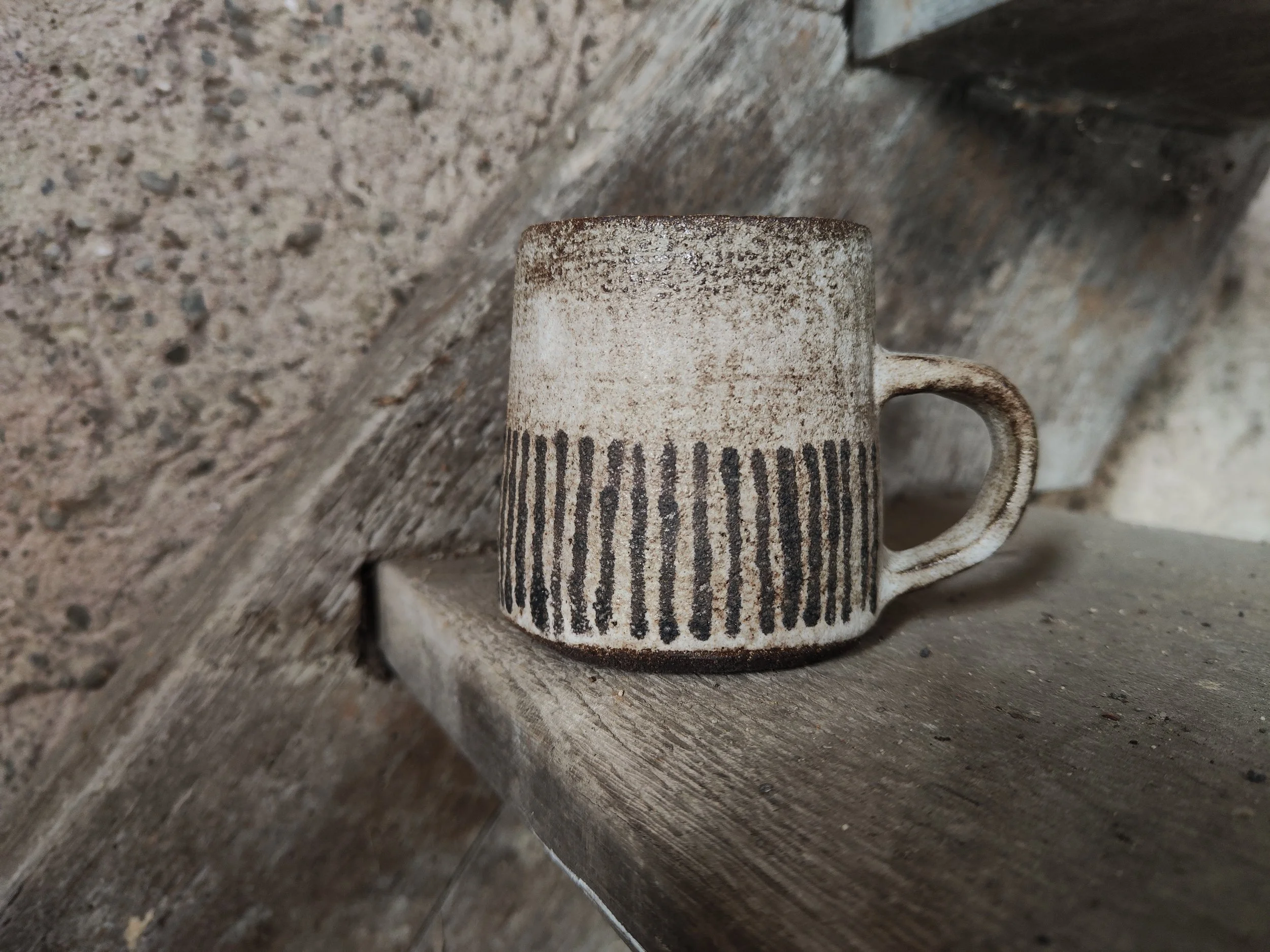 Close-up of a ceramic mug with black vertical stripes, resting on a rustic wooden surface with weathered walls in the background.