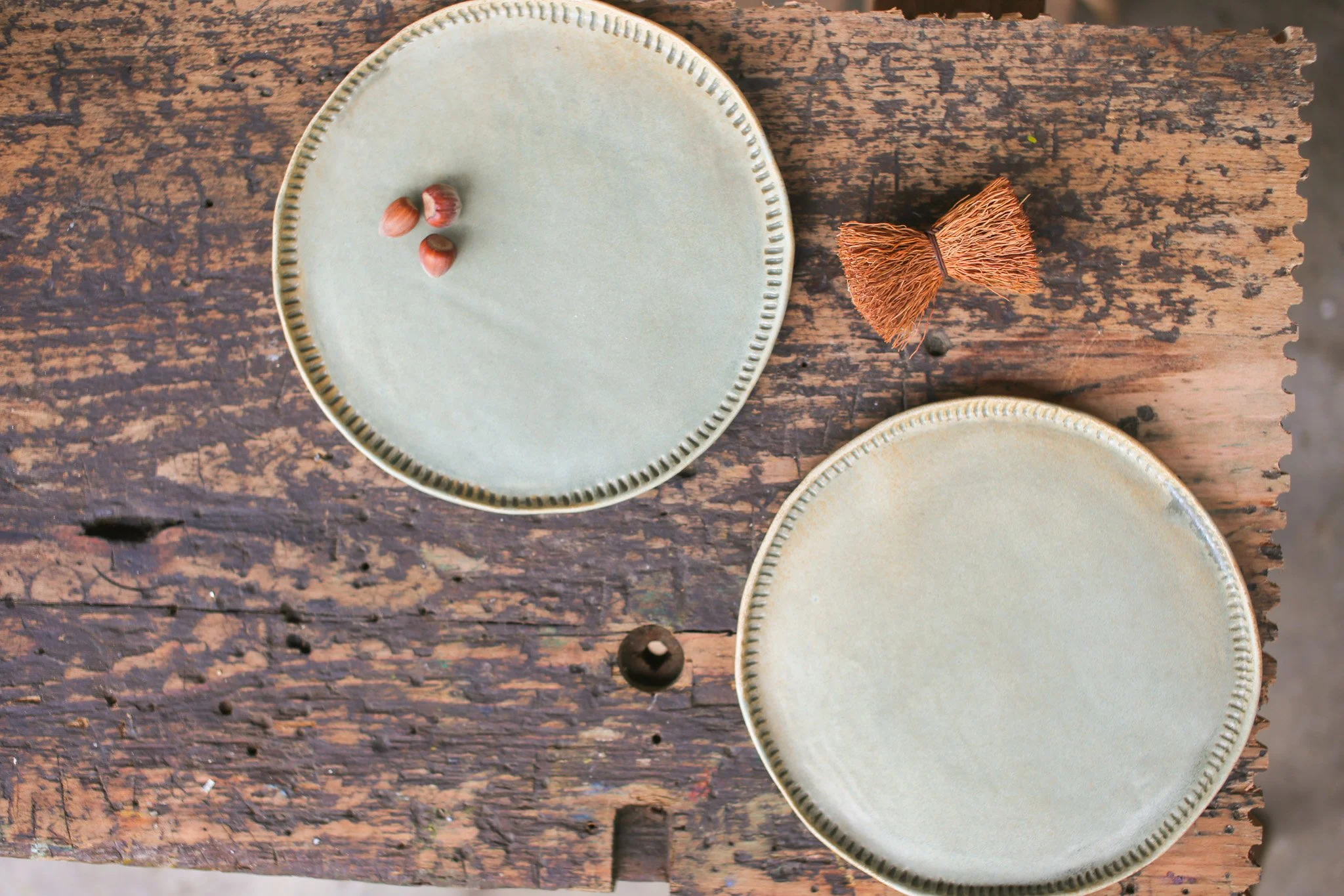 Three hazelnuts on a light-colored ceramic plate placed on a rustic wooden surface with visible grains and cracks. To the right, part of a small decorative object resembling a brown tassel or bundle of natural fibers is visible.