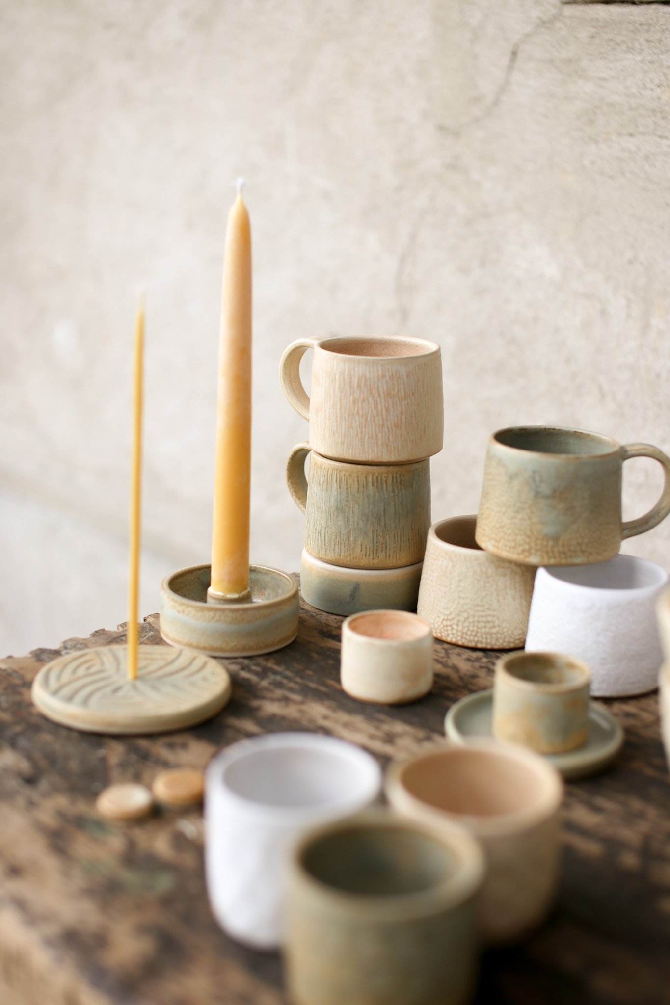 Various handmade ceramic cups and bowls arranged on a rustic wooden surface, with two tall candles in the background against a plain wall.