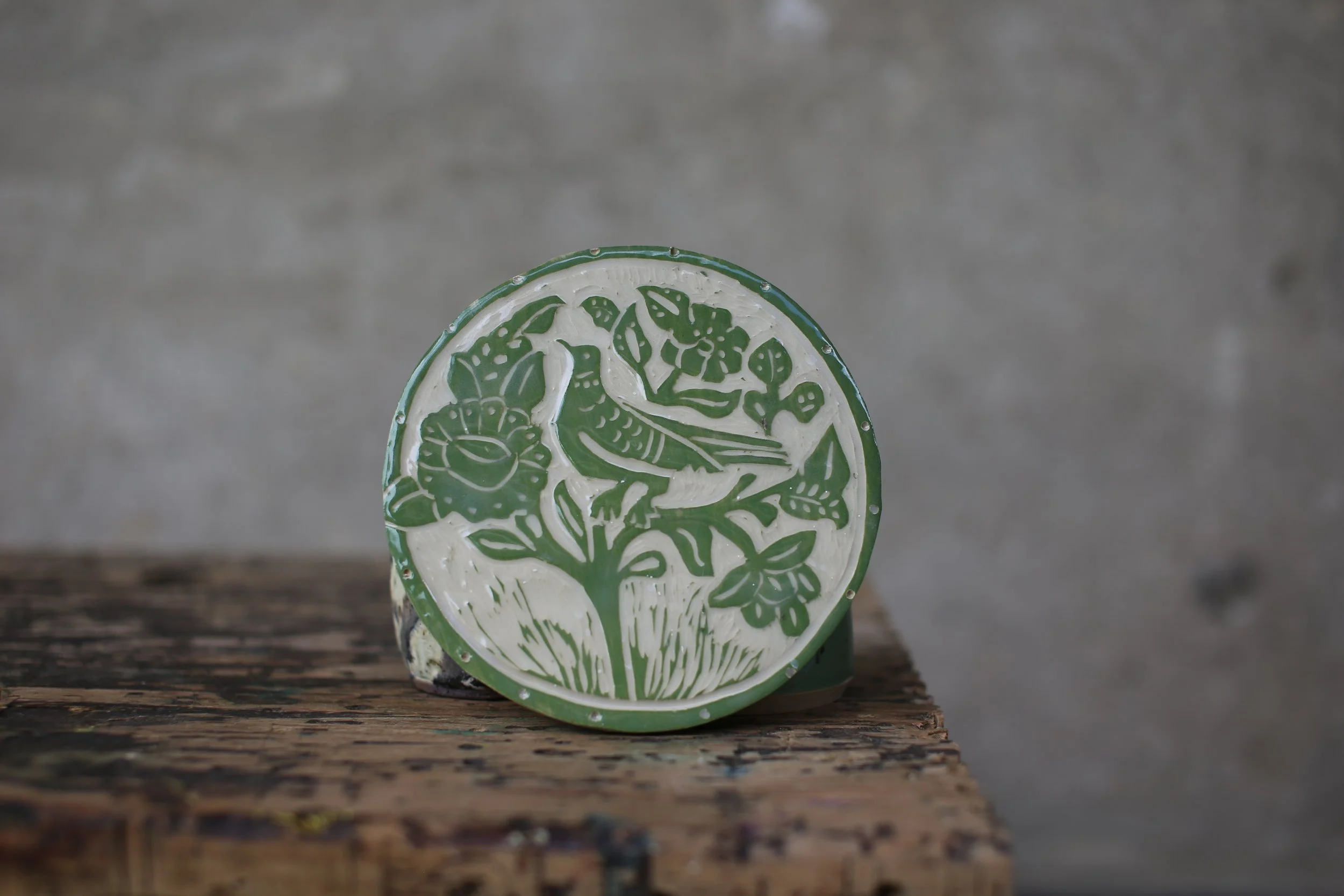 A decorative circular ceramic dish with a green and white floral pattern leaning against a gray wall on a wooden surface.