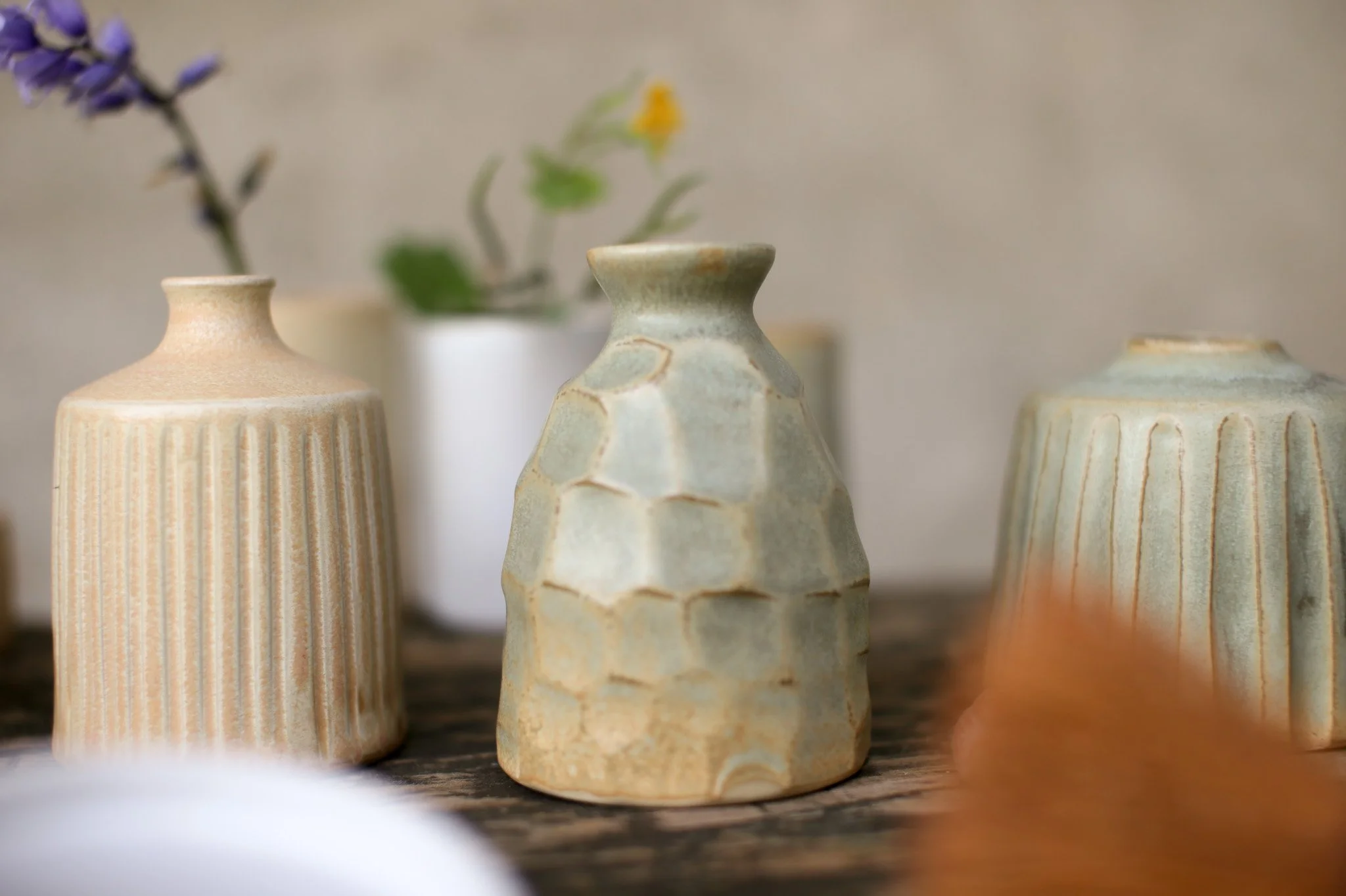 Close-up of three textured ceramic vases with floral arrangements in the background against a plain beige wall.