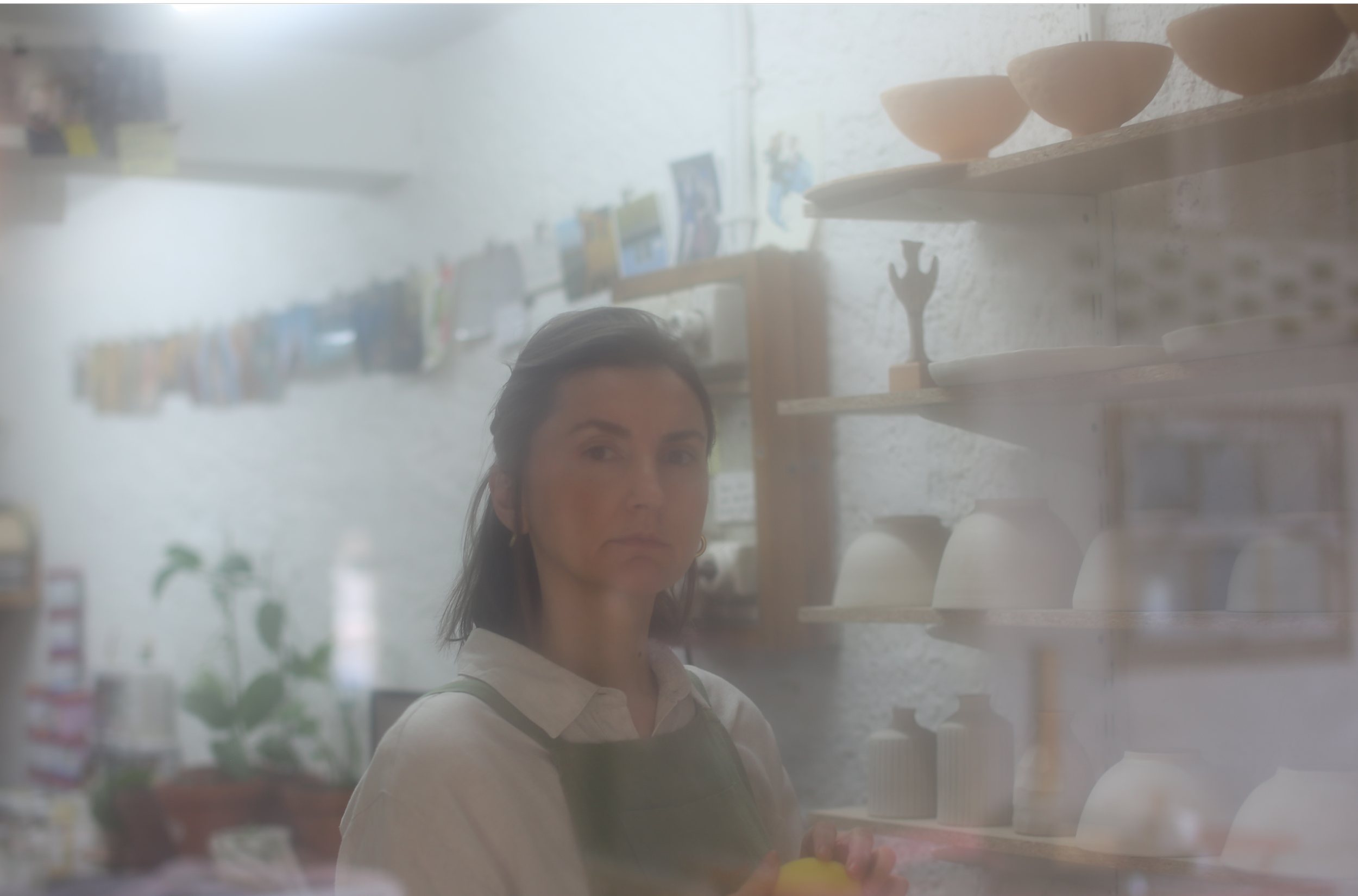 A woman with dark hair and a white shirt looking through a glass or window in a pottery workshop, with shelves containing pots visible behind her.