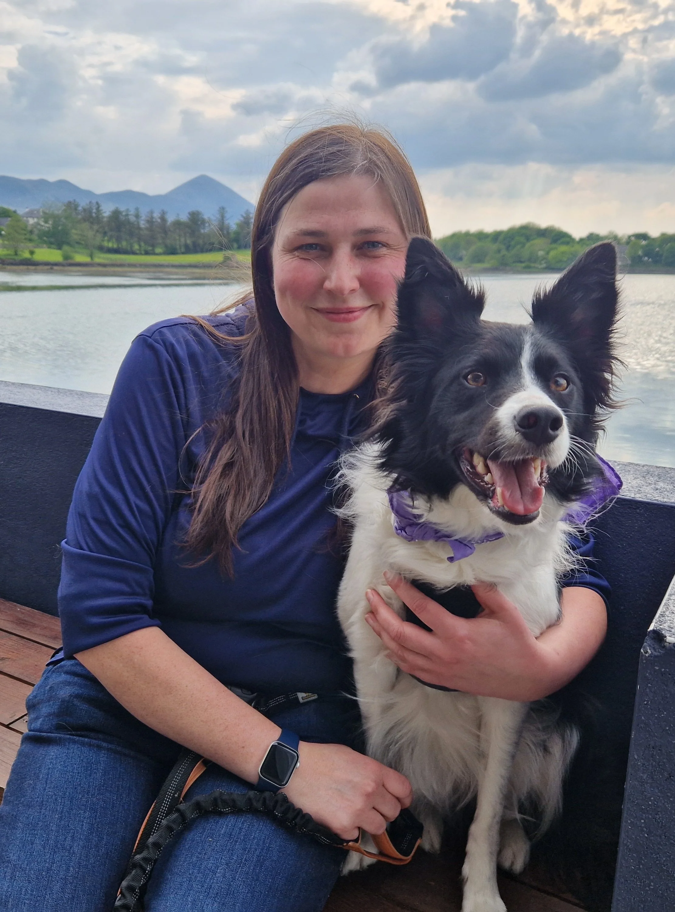 A woman with long brown hair and a blue long-sleeve shirt sitting next to a black and white Border Collie dog with a purple bandana, by a body of water with mountains and trees in the background.