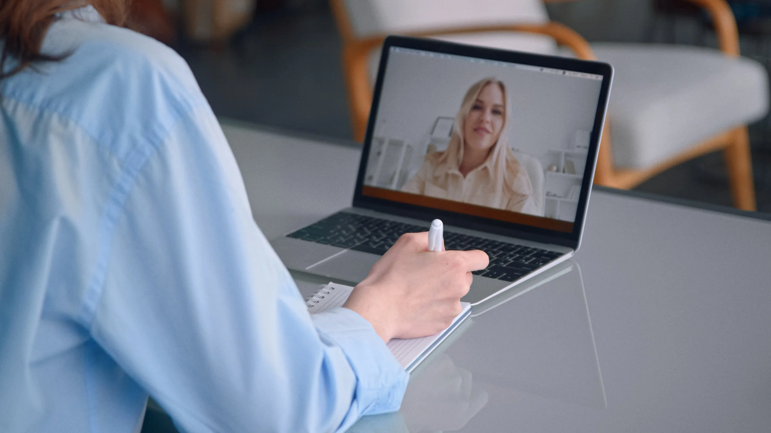 A person in a blue shirt holding a pen and taking notes on a notepad while participating in a video call on a laptop, showing a blonde woman on the screen.