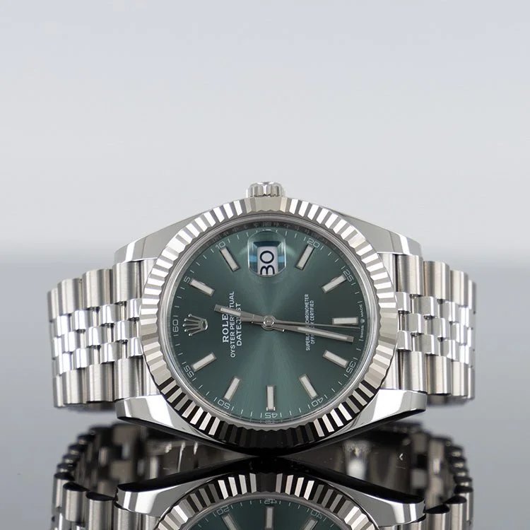 A silver Rolex wristwatch with a green face, date display at 3 o'clock, and a fluted bezel, reflected on a glossy surface.